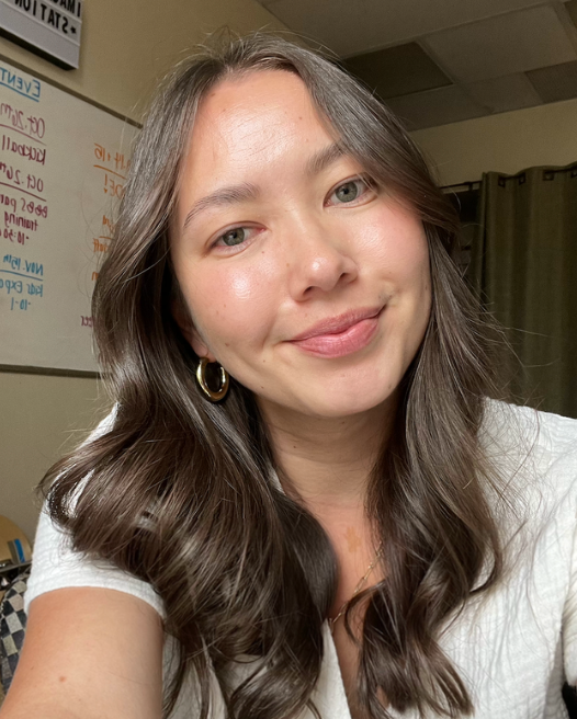 Woman with glasses smiles while sitting on a bench in a park; wearing a white jacket over a black top.