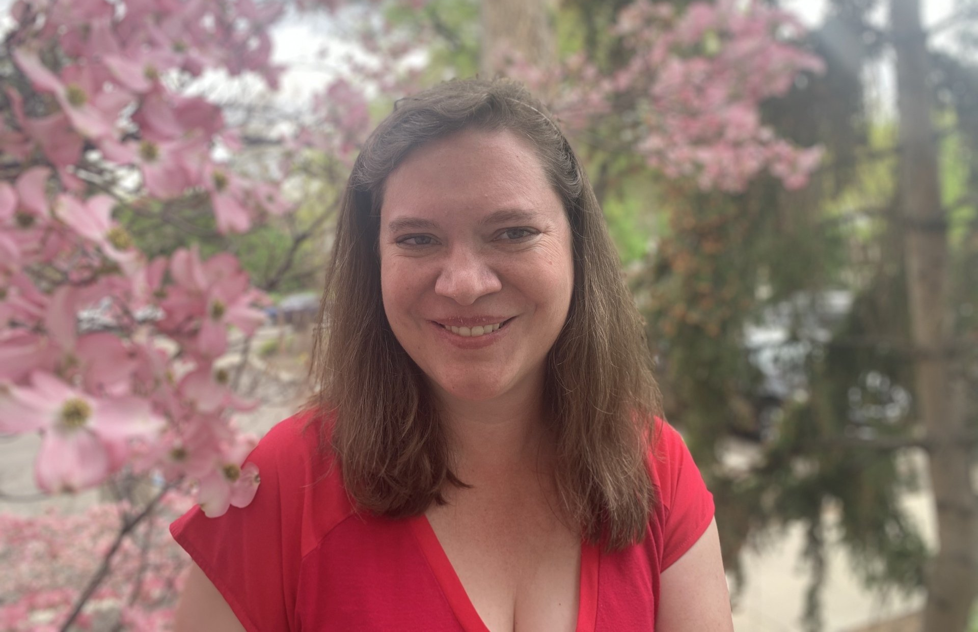 A woman in a red shirt is smiling in front of a tree with pink flowers.