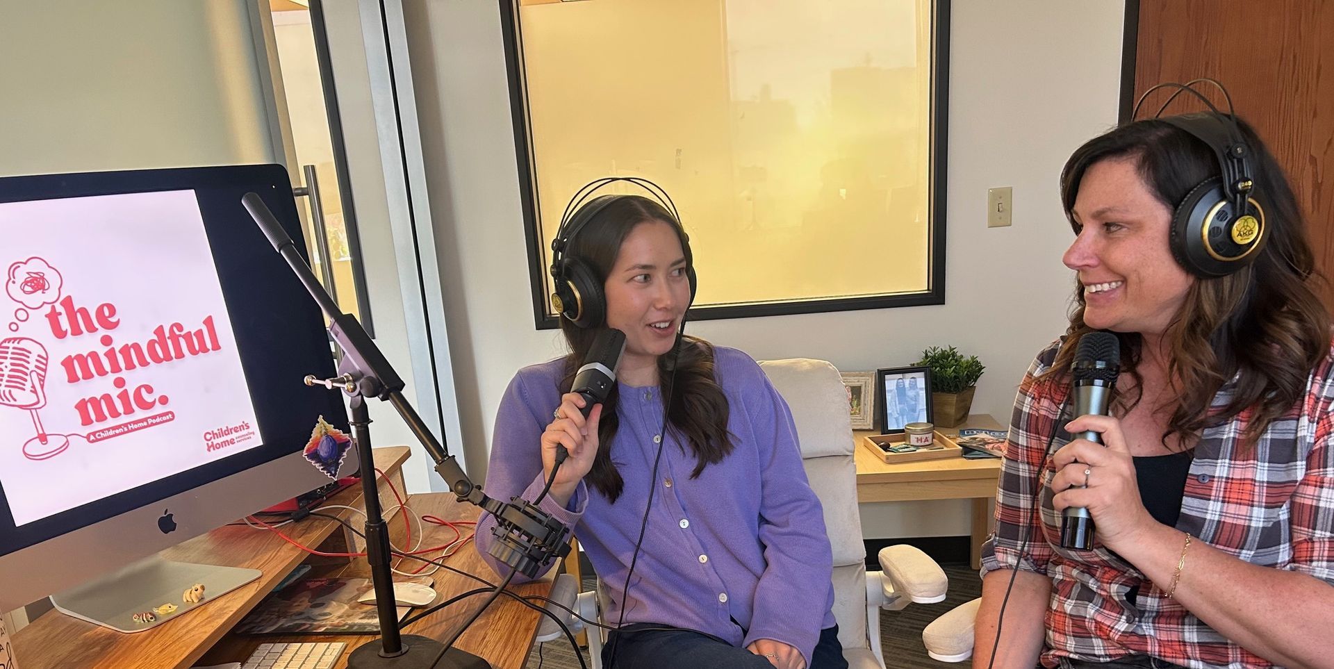 Two women in a podcast studio, speaking into microphones. One wears a purple shirt, the other a plaid shirt.
