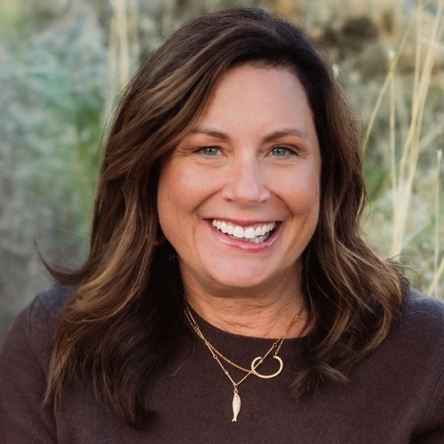 Woman with brown hair, smiling, wearing a brown shirt and gold necklace, outdoors.