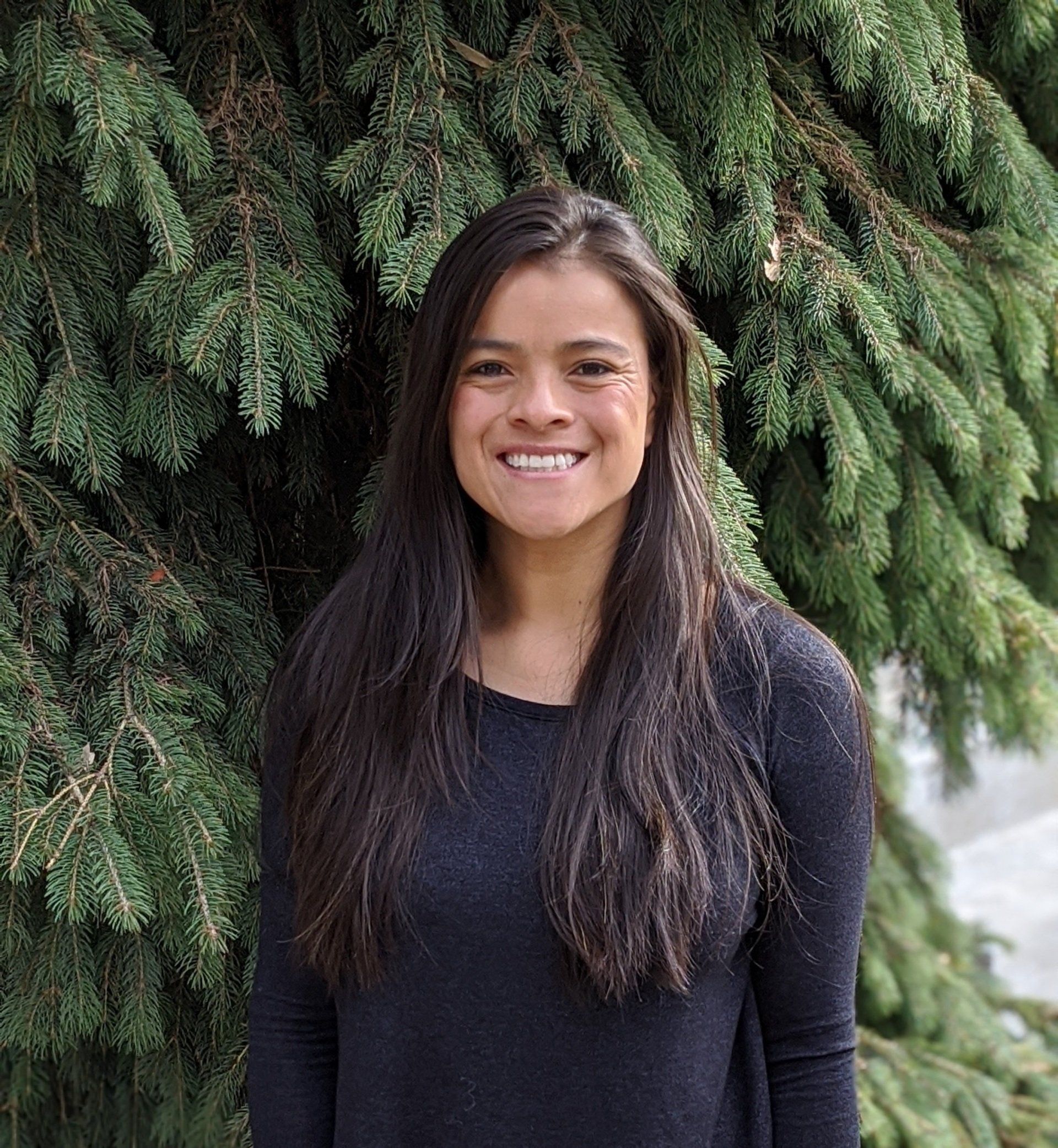 A woman in a black shirt is smiling in front of a pine tree.