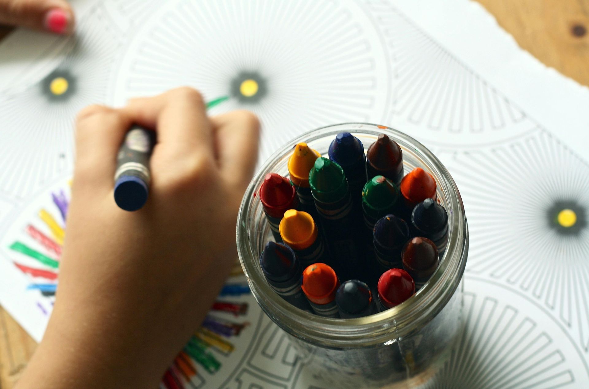 A hand coloring a drawing with a blue crayon, surrounded by a jar of colorful crayons.