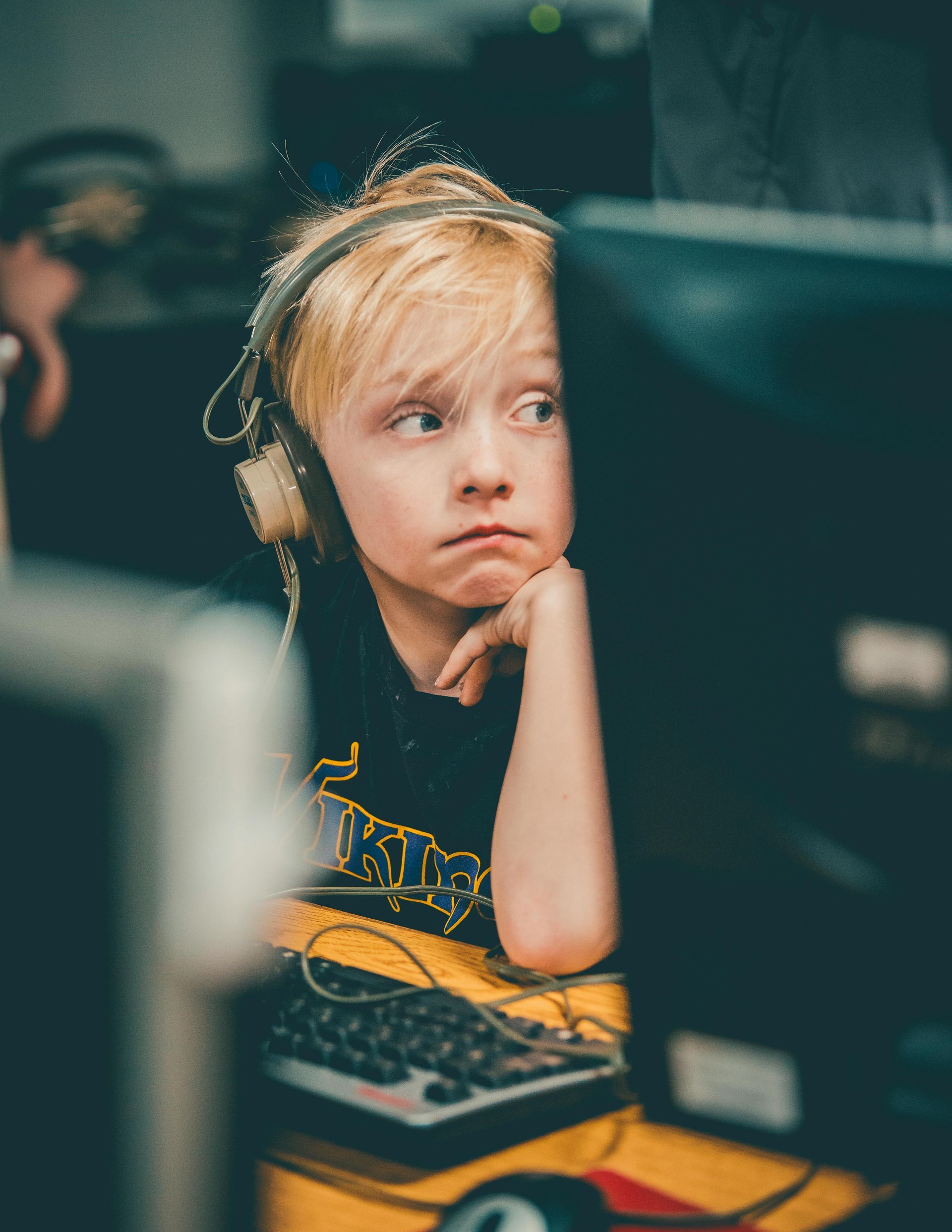 Blond child wearing headphones, looking off to the side with a pensive expression while sitting at a computer.