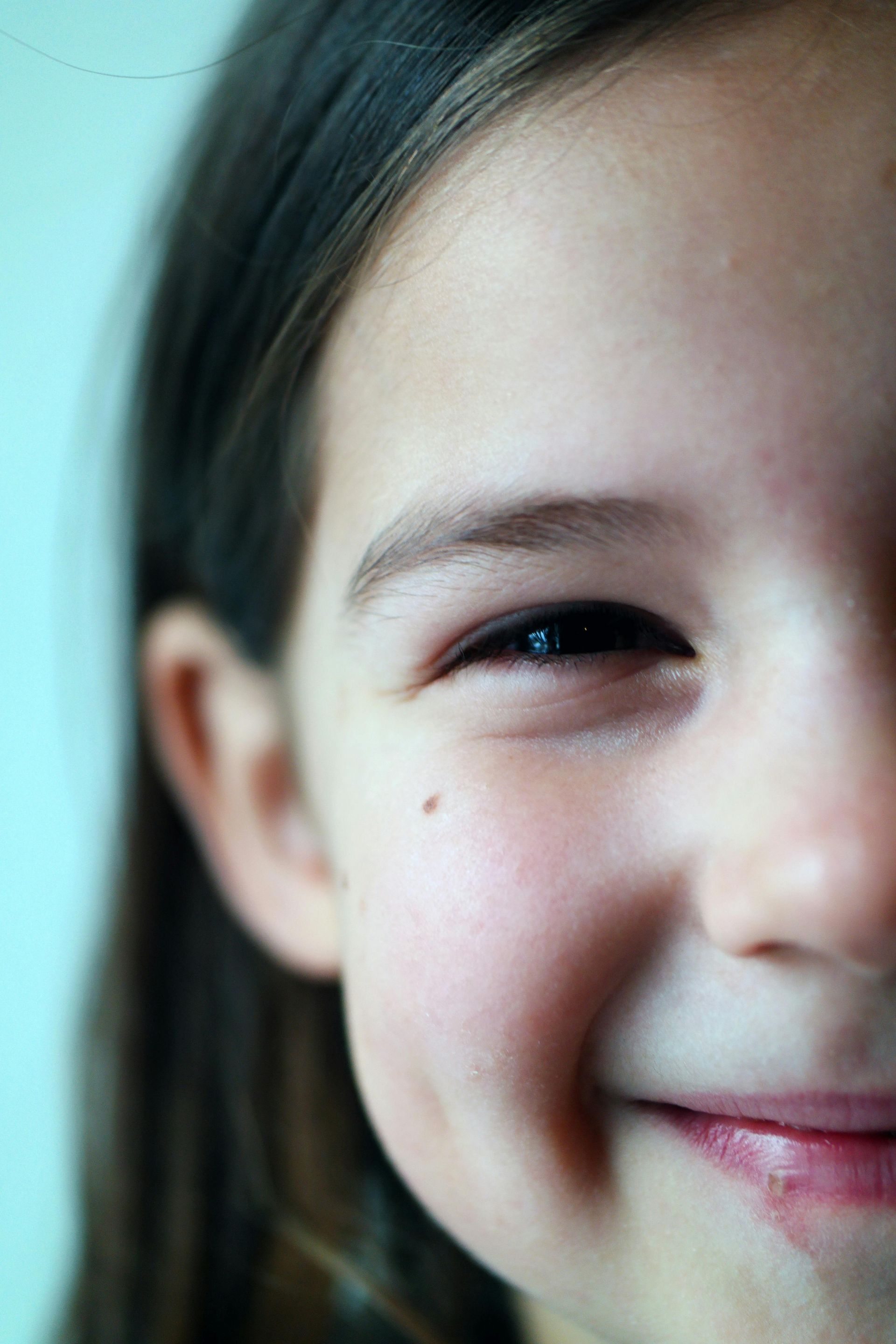 Close-up of a smiling child with dark hair, a mole on their cheek, and a visible dimple.