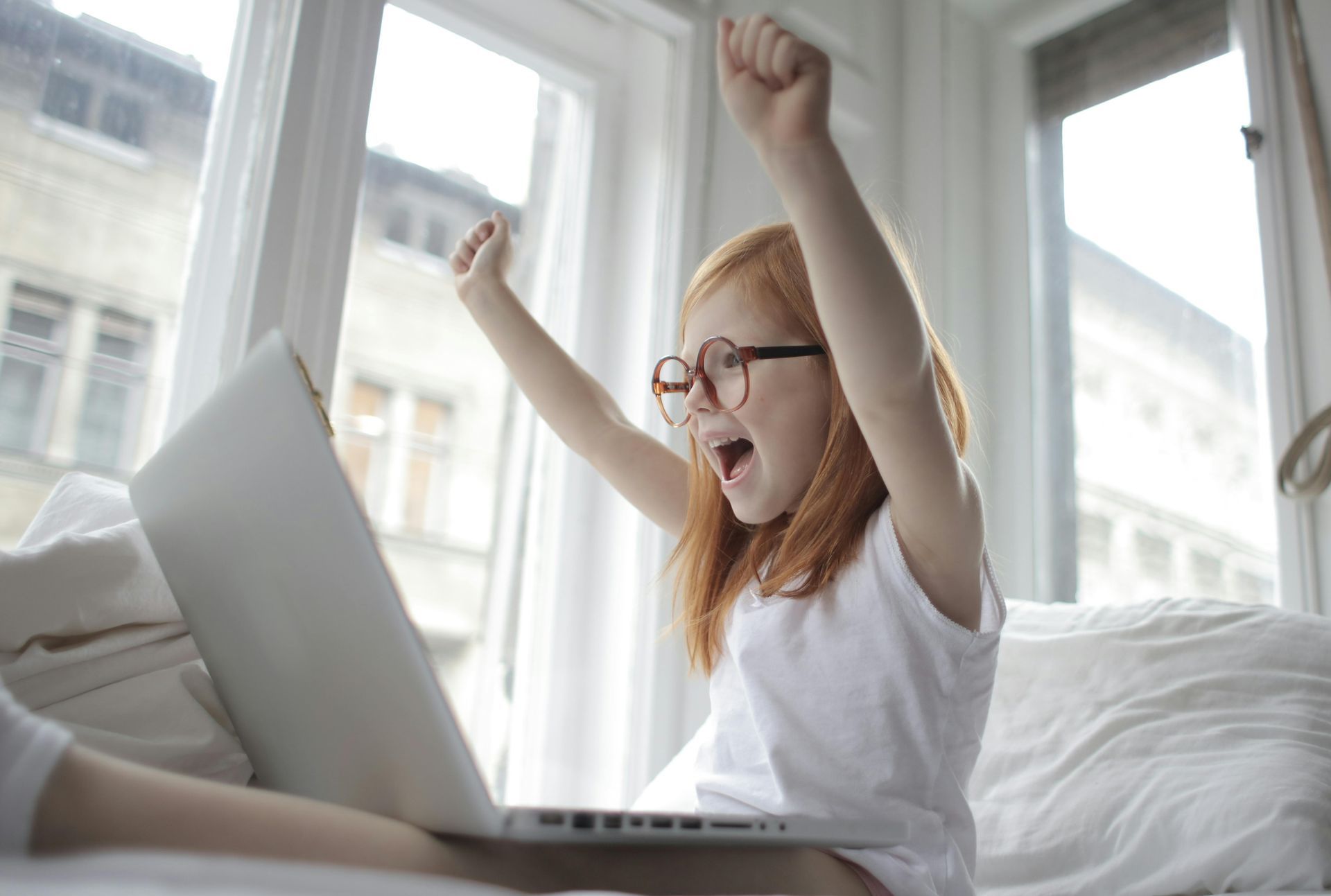 Girl with red hair and glasses cheers in front of a laptop, arms raised. Bright, natural light floods the room.