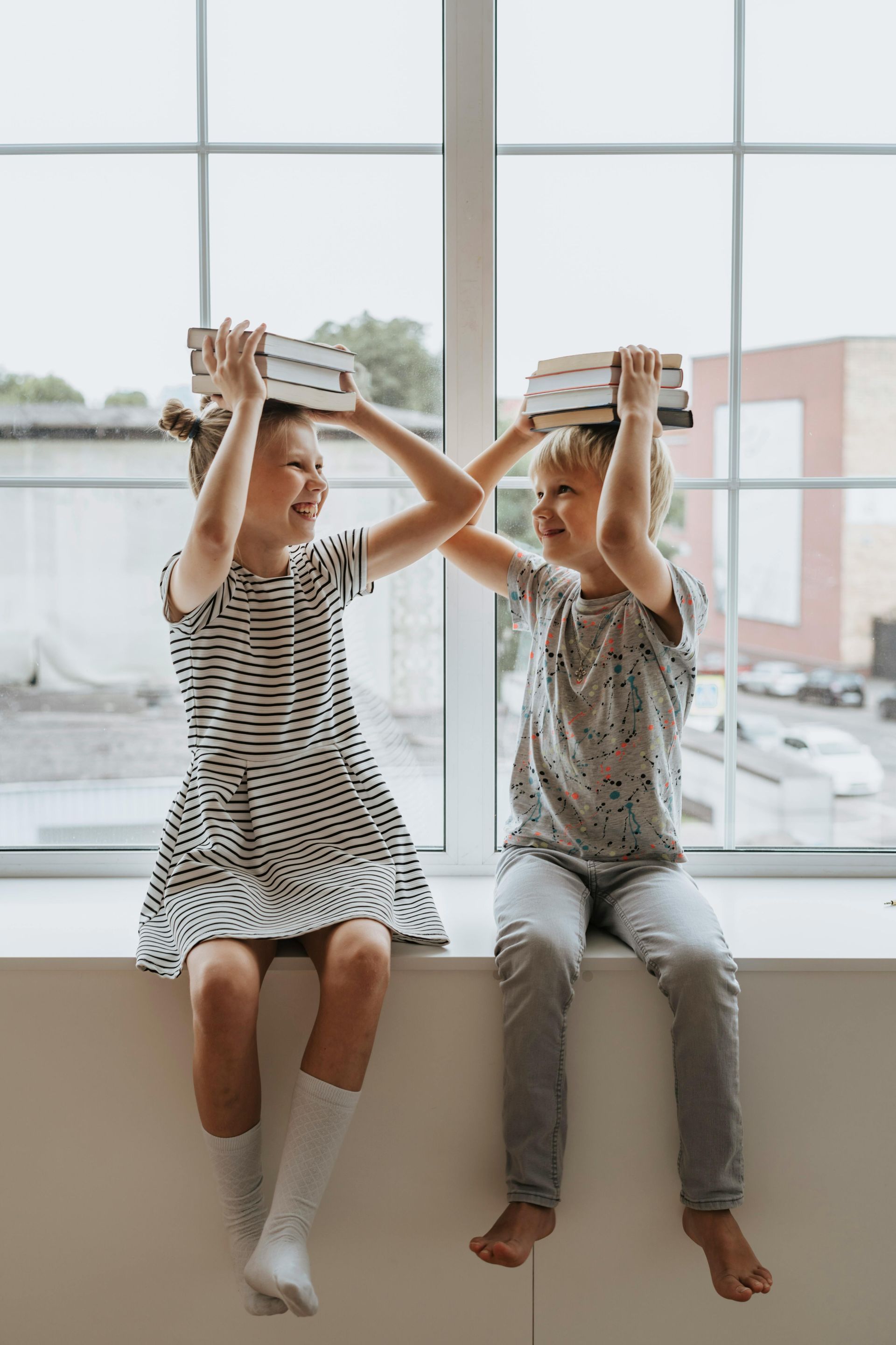 Two children balancing books on their heads while sitting on a windowsill, smiling.