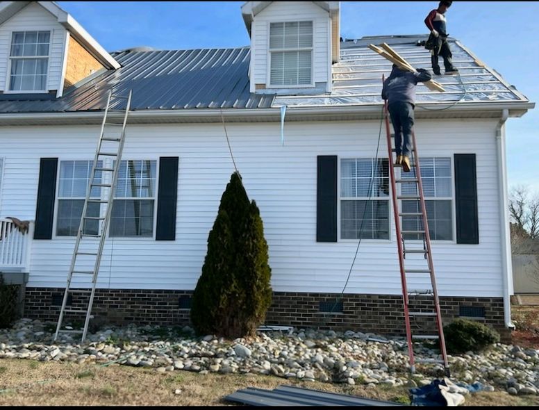 Roofers installing metal roofing on a white house with black shutters. Ladders and stacked wood present.