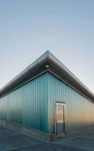Triangular building with teal glass walls, metal trim, and a door, against a clear blue sky.