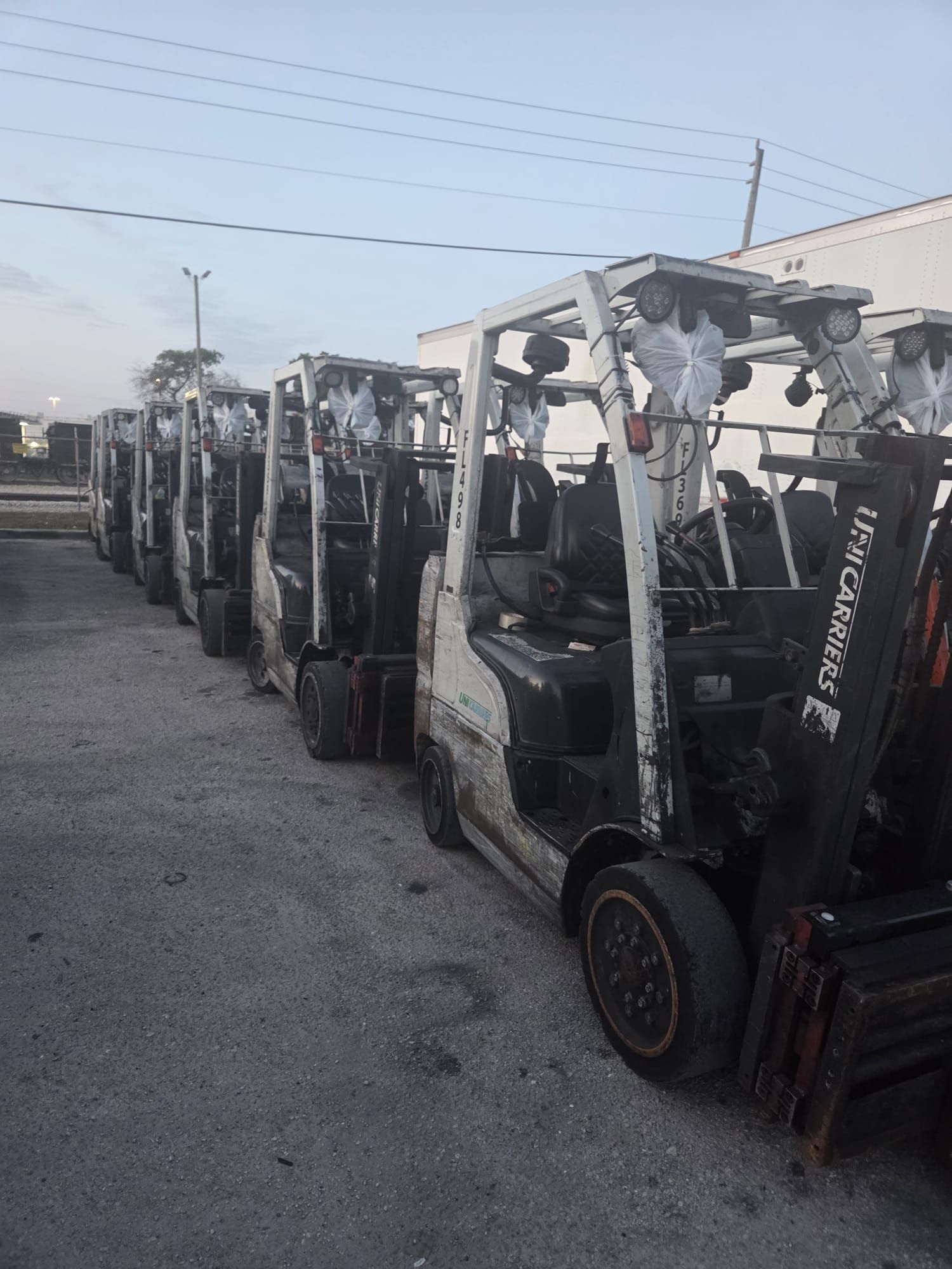 Row of white forklifts parked on concrete, under a cloudy sky.