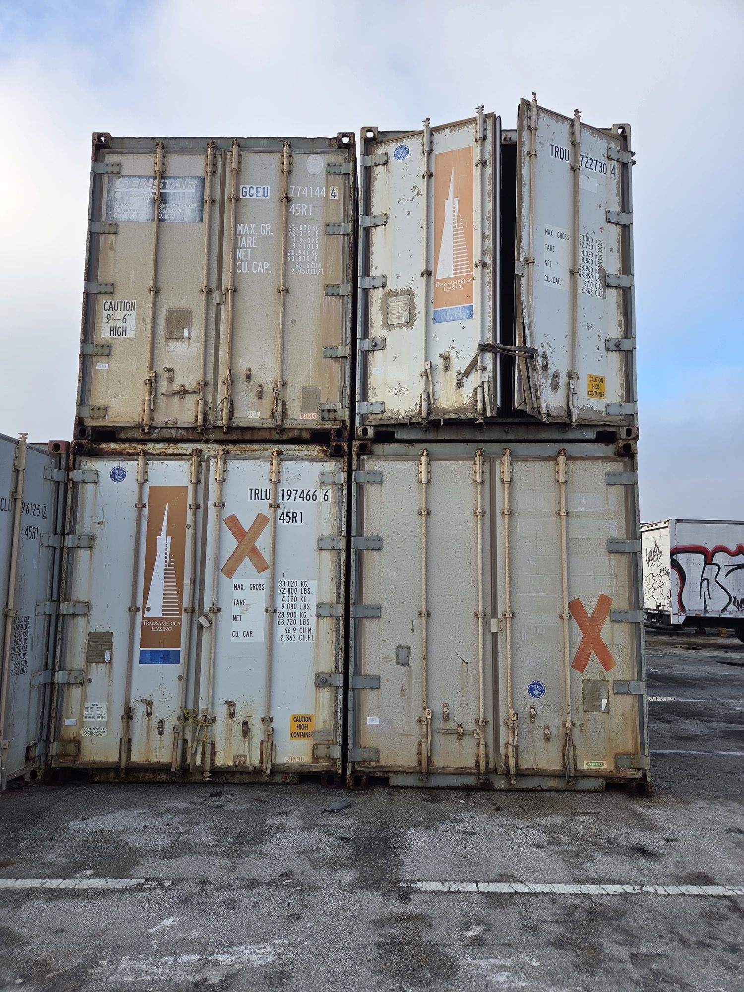 Four stacked shipping containers, weathered gray, with one door open, against a cloudy sky.