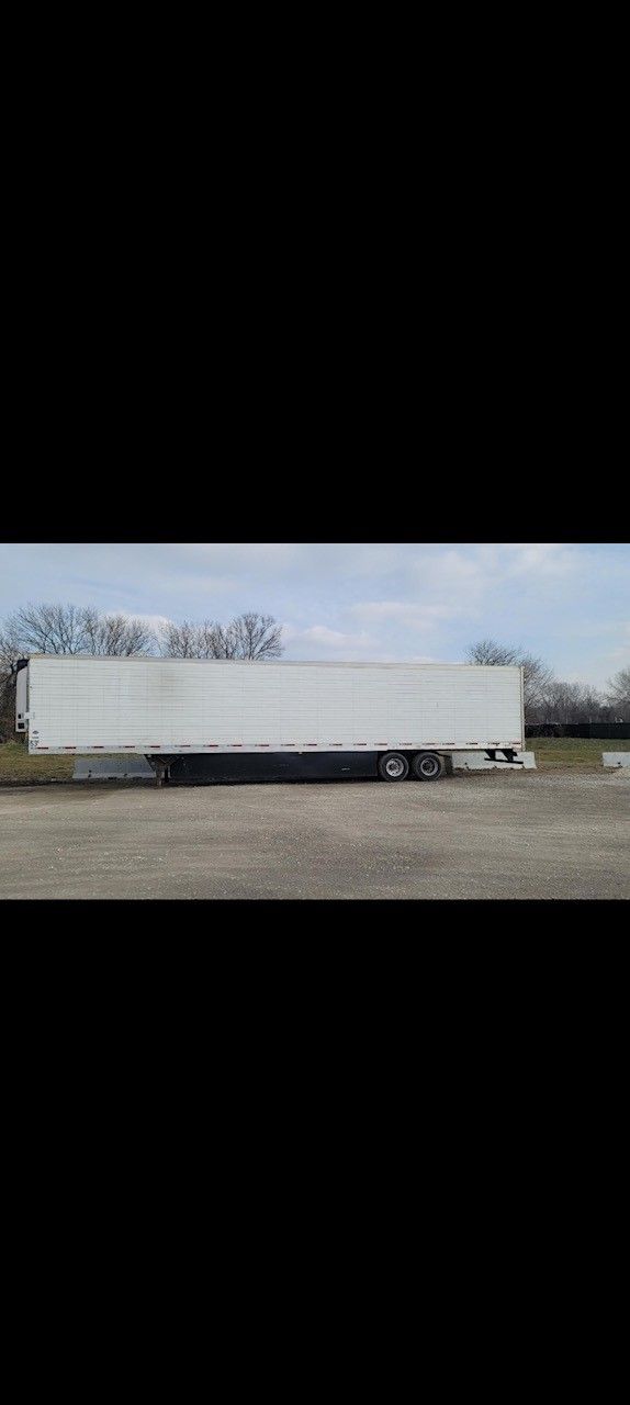 A white semi-trailer parked on gravel with a backdrop of trees and a cloudy sky.