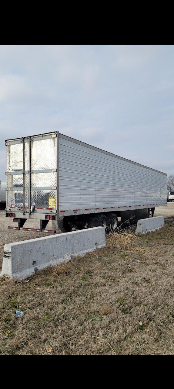 A large, gray semi-trailer parked on a patch of brown grass next to concrete barriers.