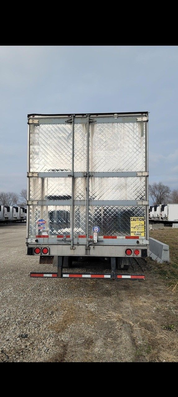 Rear view of a silver cargo trailer in a gravel lot under a cloudy sky.