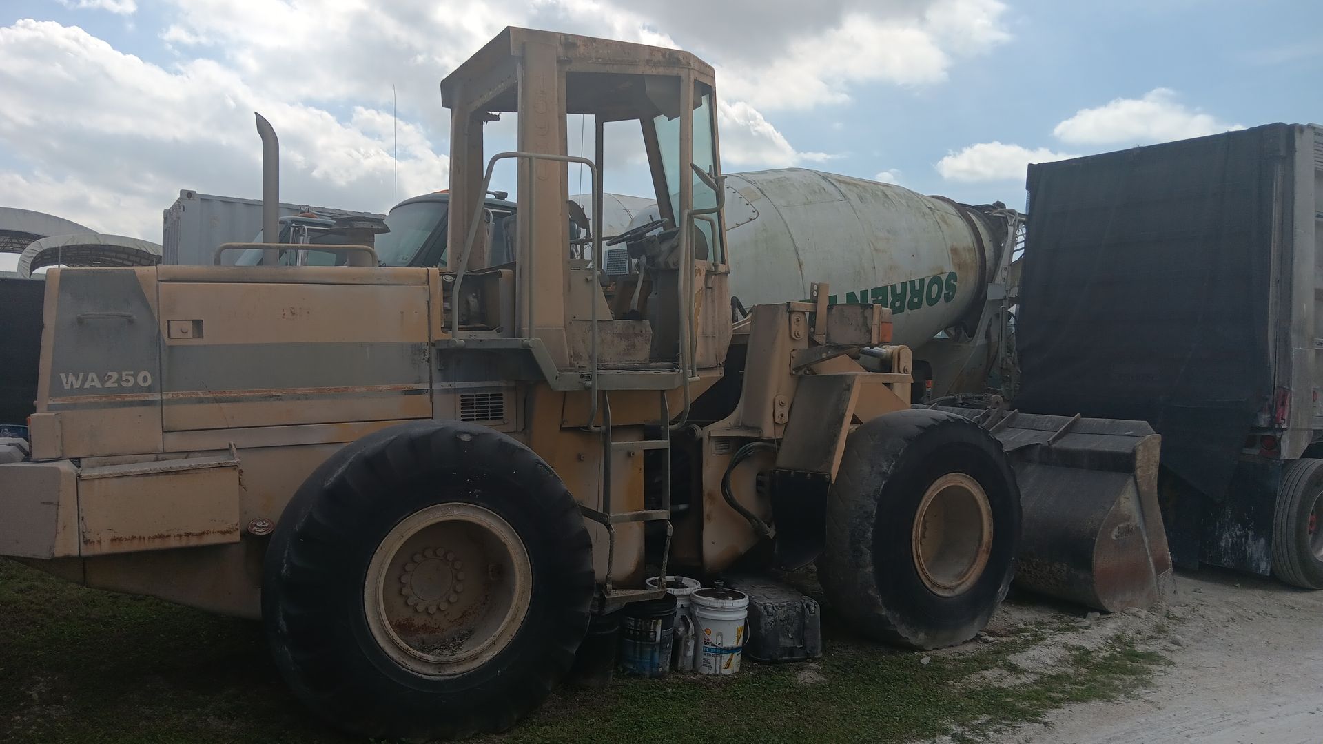 Yellow front-end loader next to a cement mixer and a black trailer on a cloudy day.
