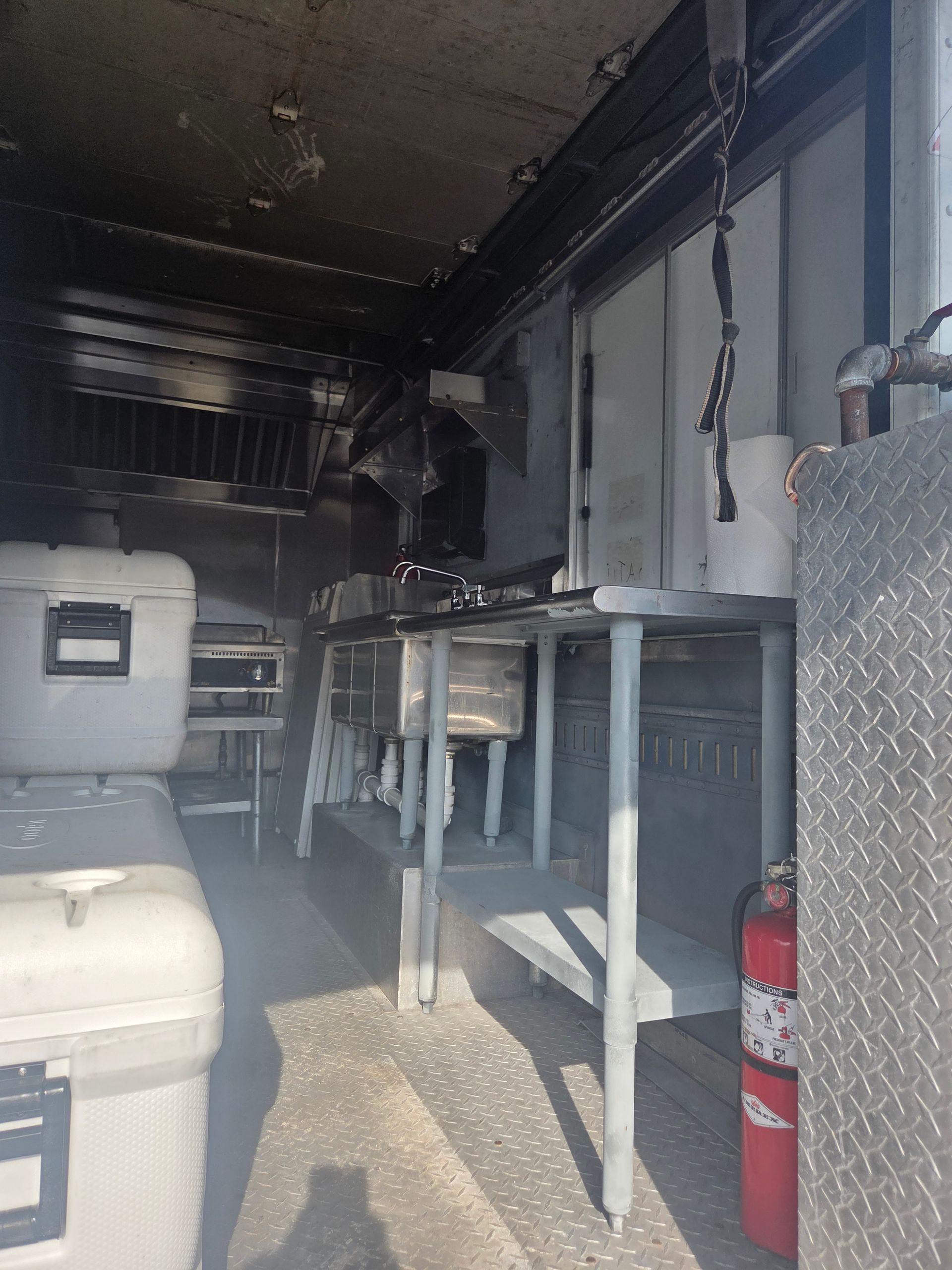 Interior of a food truck, damage visible. Stainless steel counters, fire extinguisher, and coolers. Smoke present.