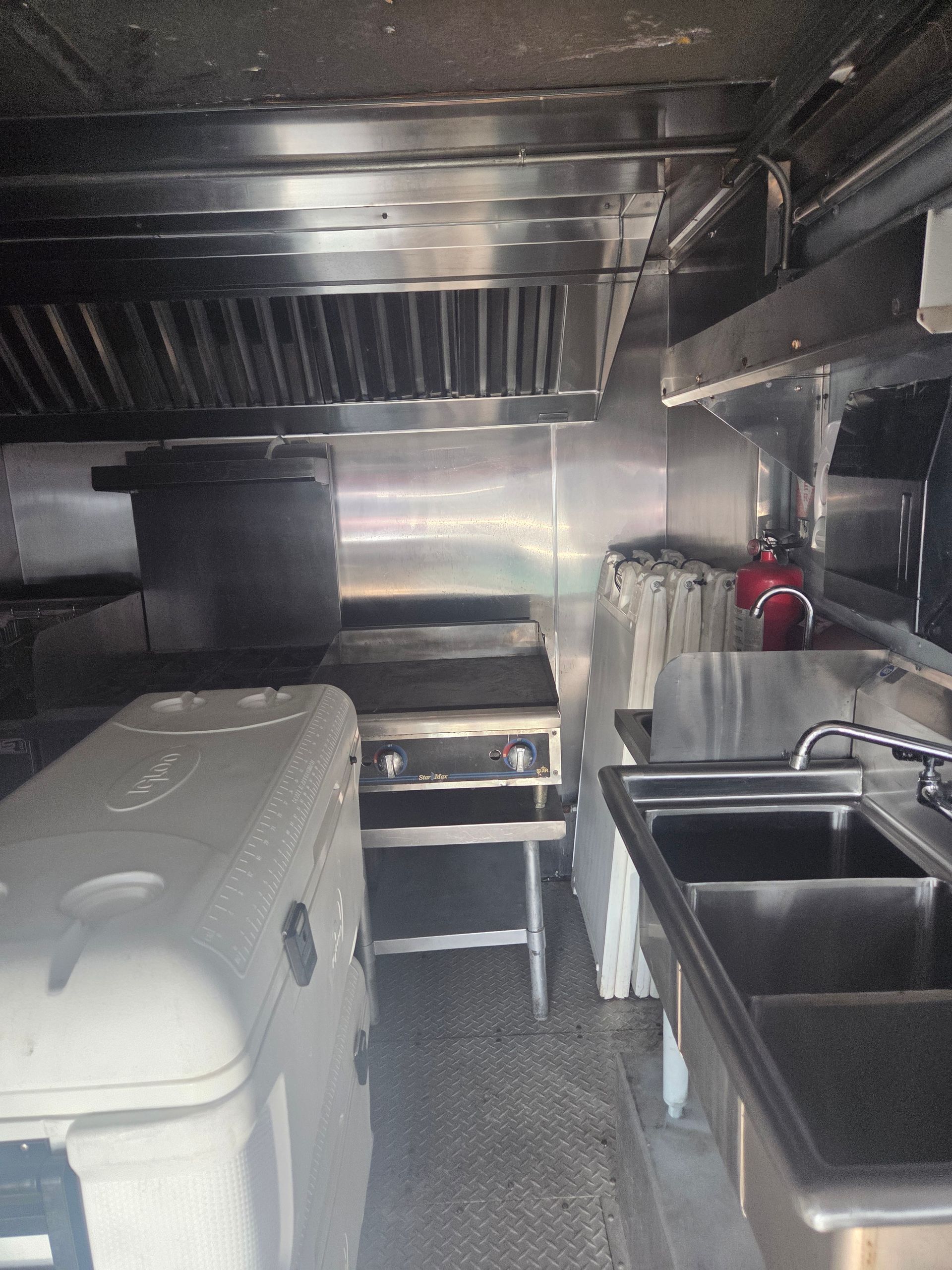 Interior of a food truck. Stainless steel appliances, grill, sinks, cooler, and hood.
