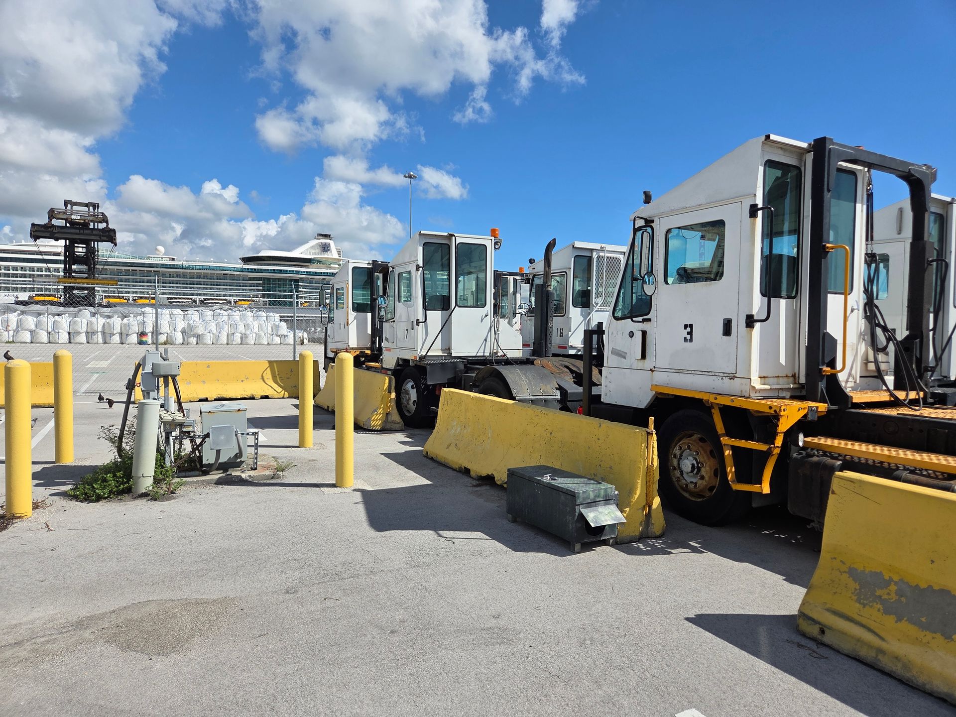 White industrial trucks parked behind yellow concrete barriers at a port, blue sky with cruise ships in the background.