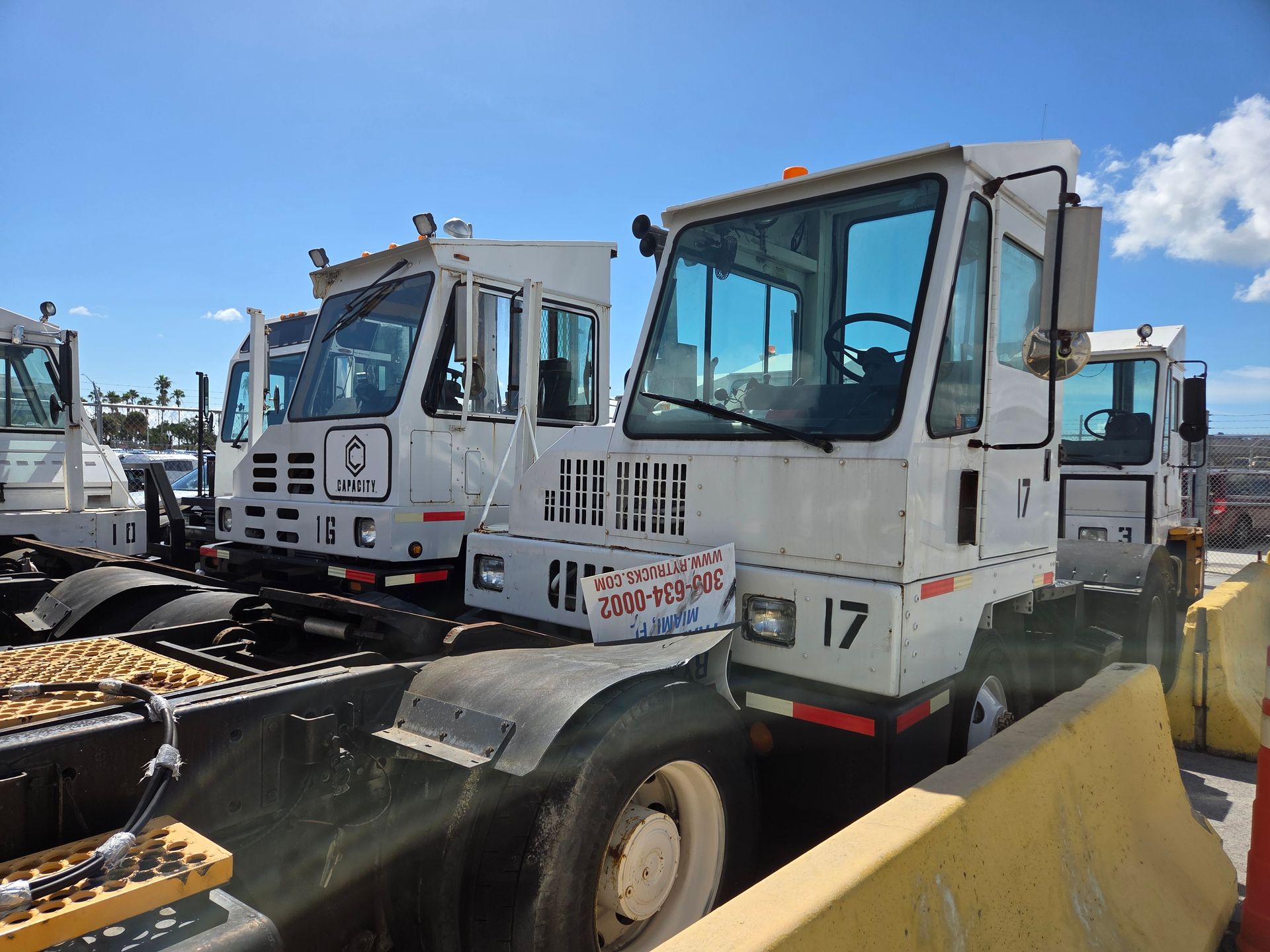 White terminal tractors parked outdoors on a sunny day, numbered 17, with safety barriers.