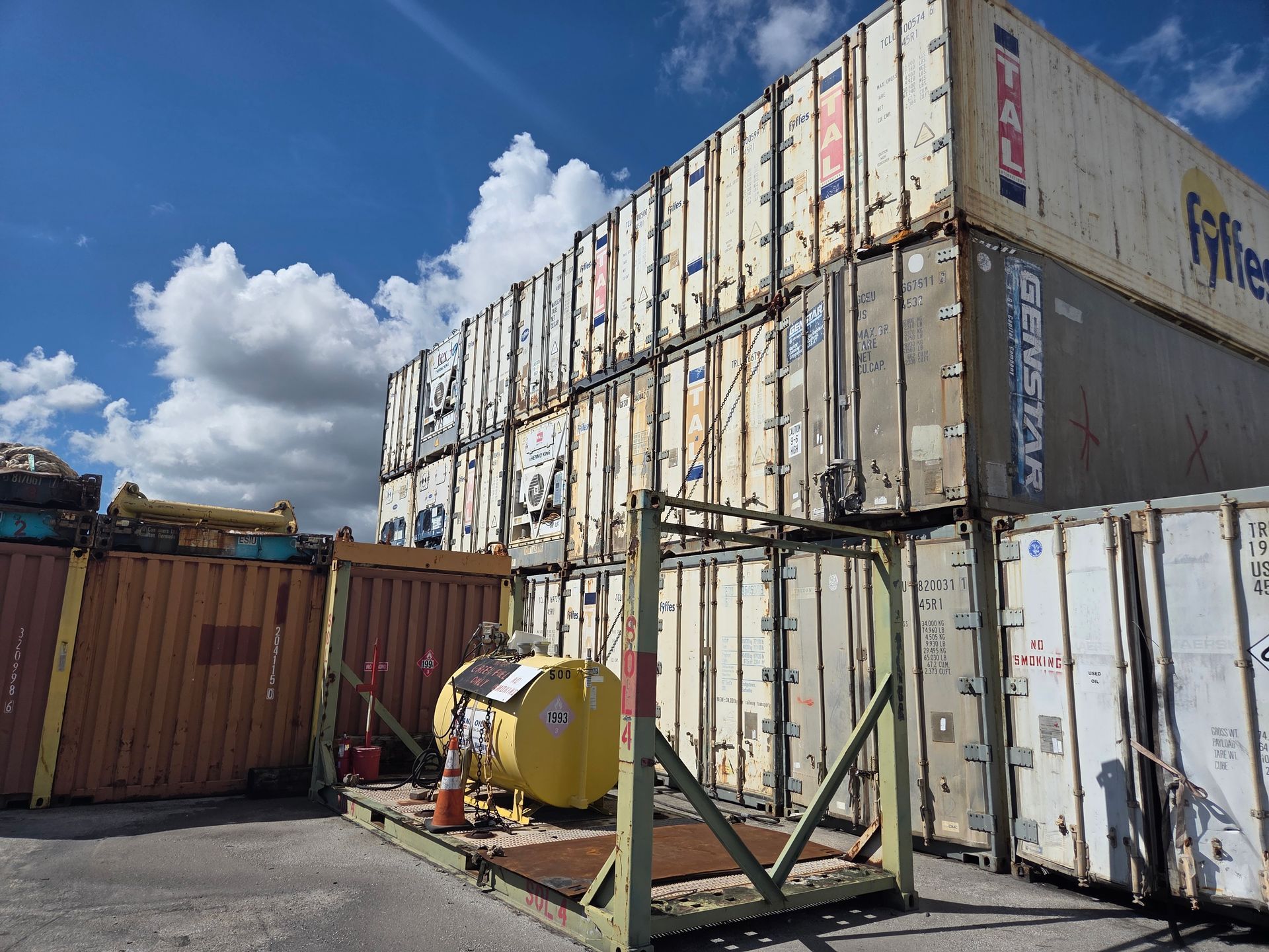 Stack of shipping containers with a yellow tank on a metal stand in an outdoor lot, under a cloudy blue sky.