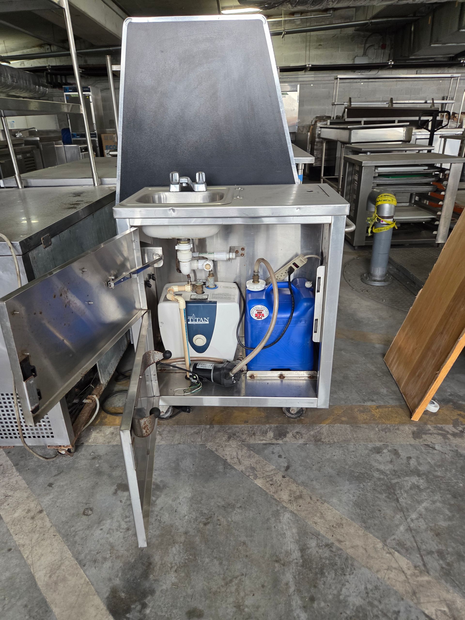 Stainless steel commercial sink cabinet with open door, revealing plumbing and machinery. Rolls on wheels.