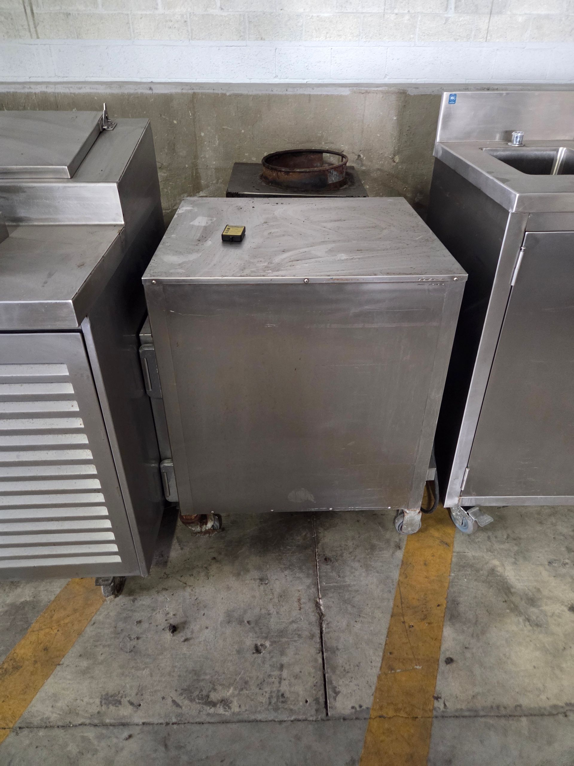 Stainless steel commercial refrigerator with other steel equipment against a wall on a concrete floor.