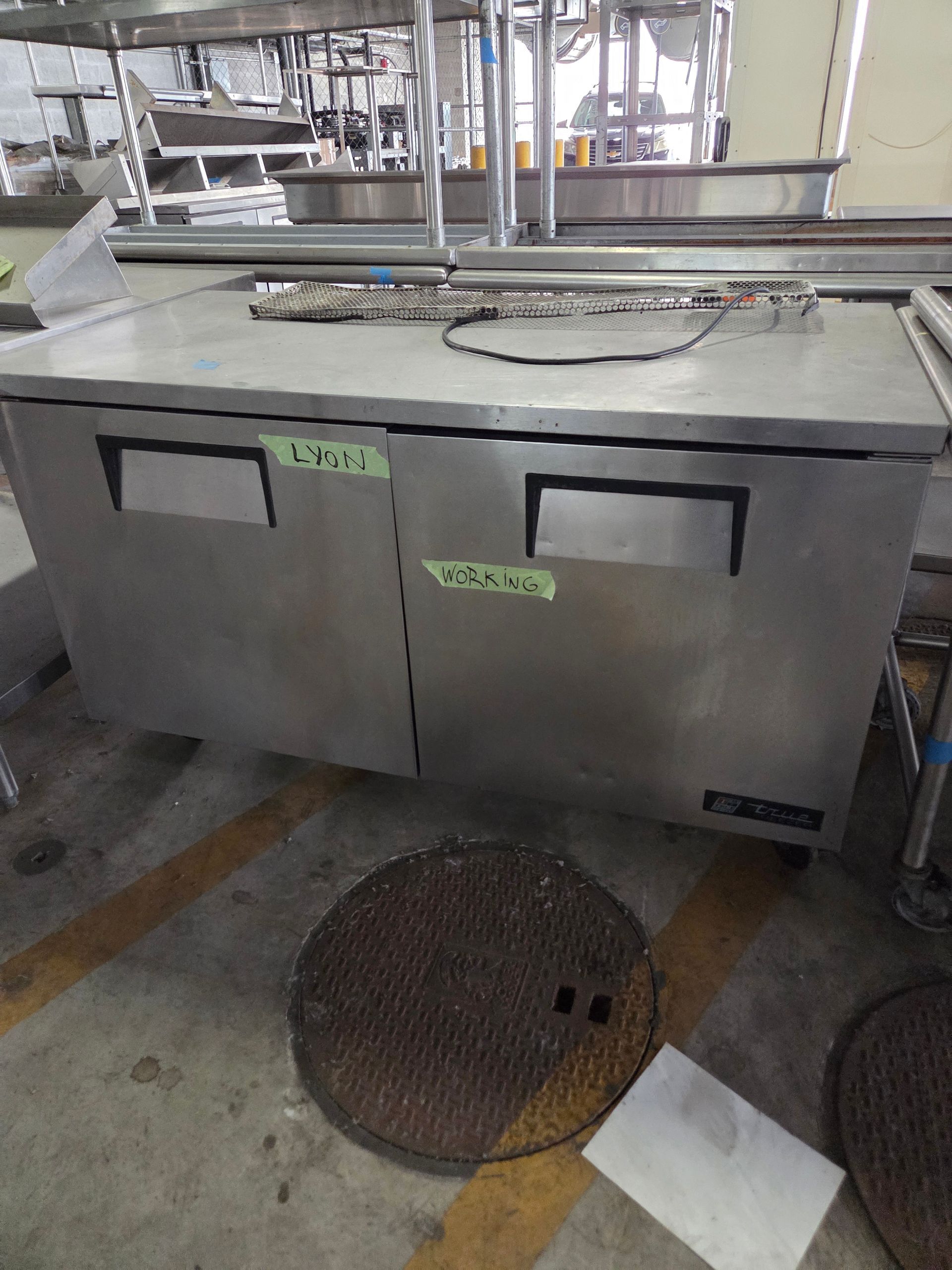 Stainless steel two-door refrigerator in a kitchen setting, with green labels on doors and a manhole cover nearby.