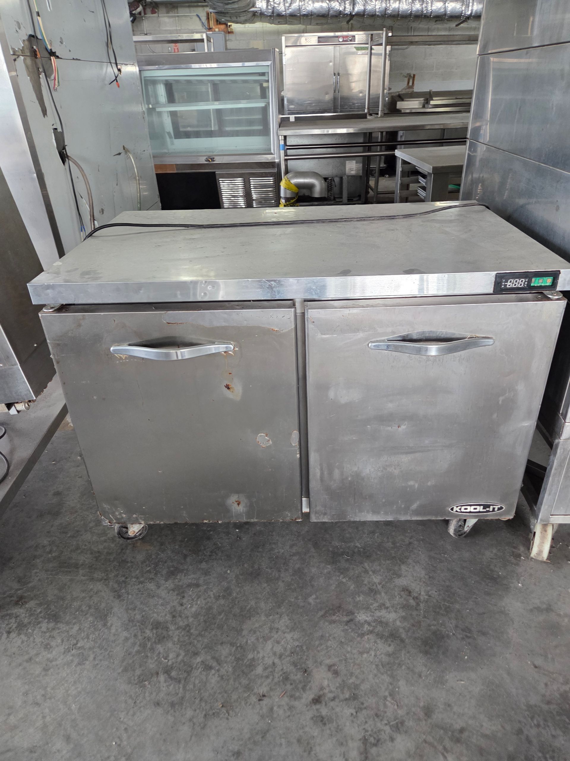 Stainless steel two-door commercial refrigerator with handles and wheels, in a kitchen setting.