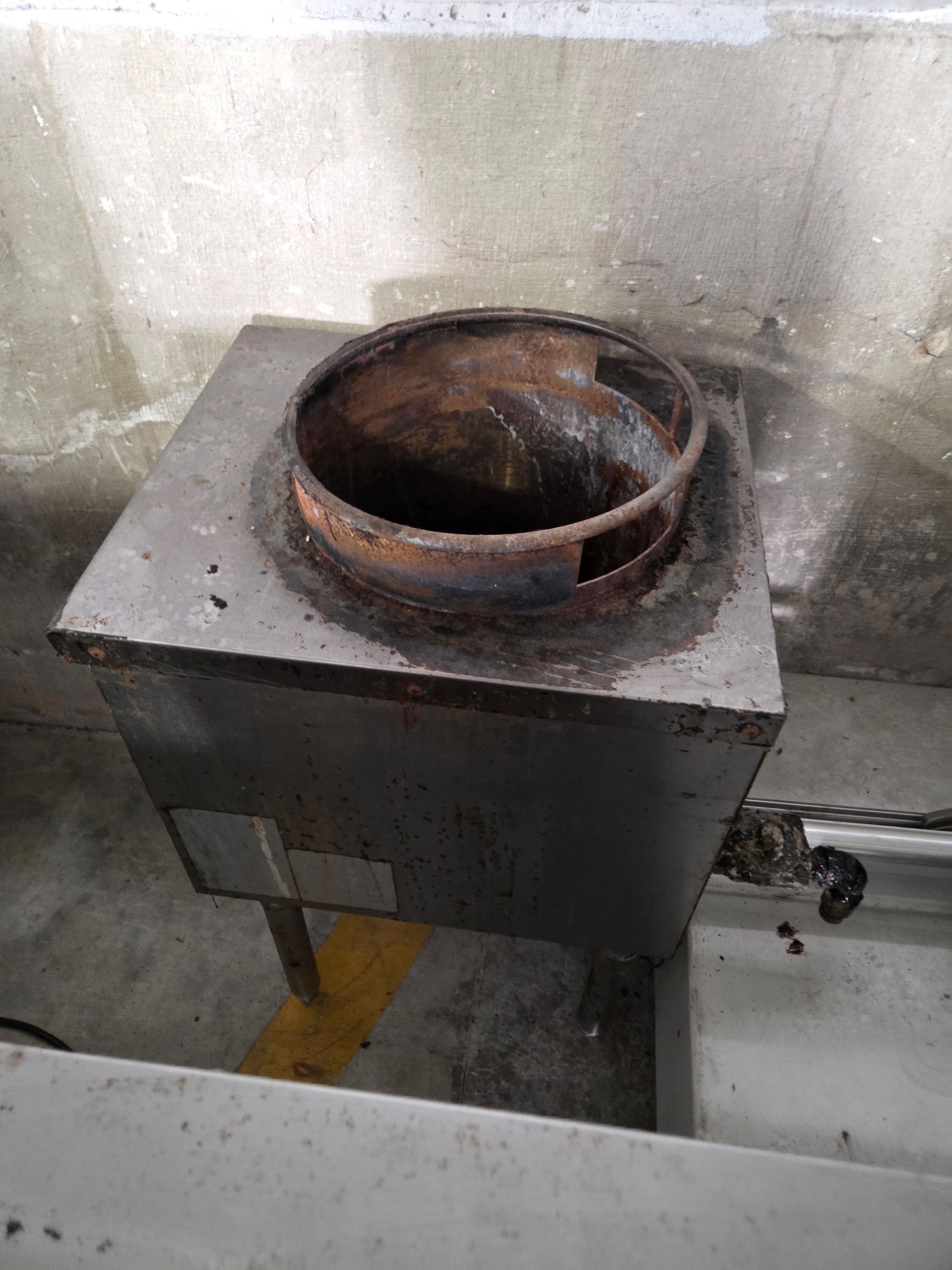 A rusty, metal wok-style stove on a gray metal box with a concrete wall background.