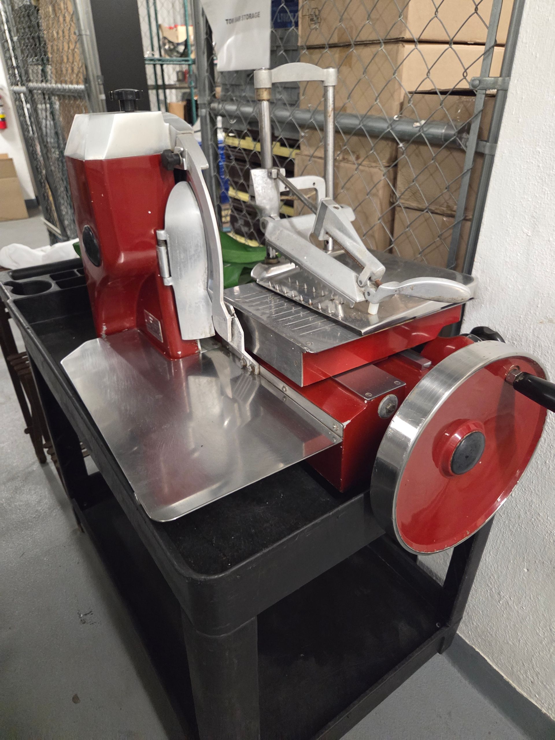 Red commercial meat slicer on black cart in a storage area.