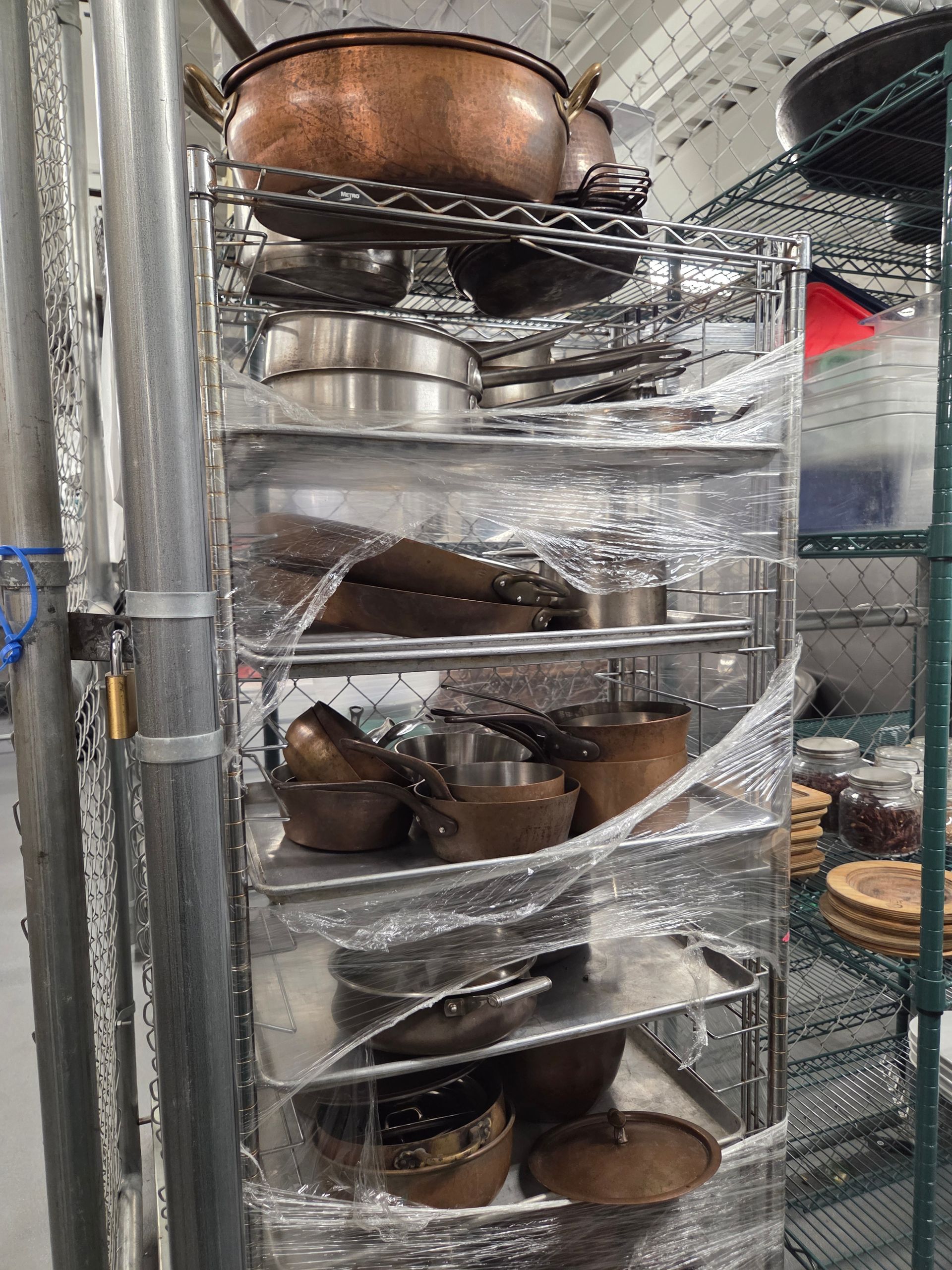 Metal shelving unit, wrapped in plastic, holding various pots and pans in a kitchen.