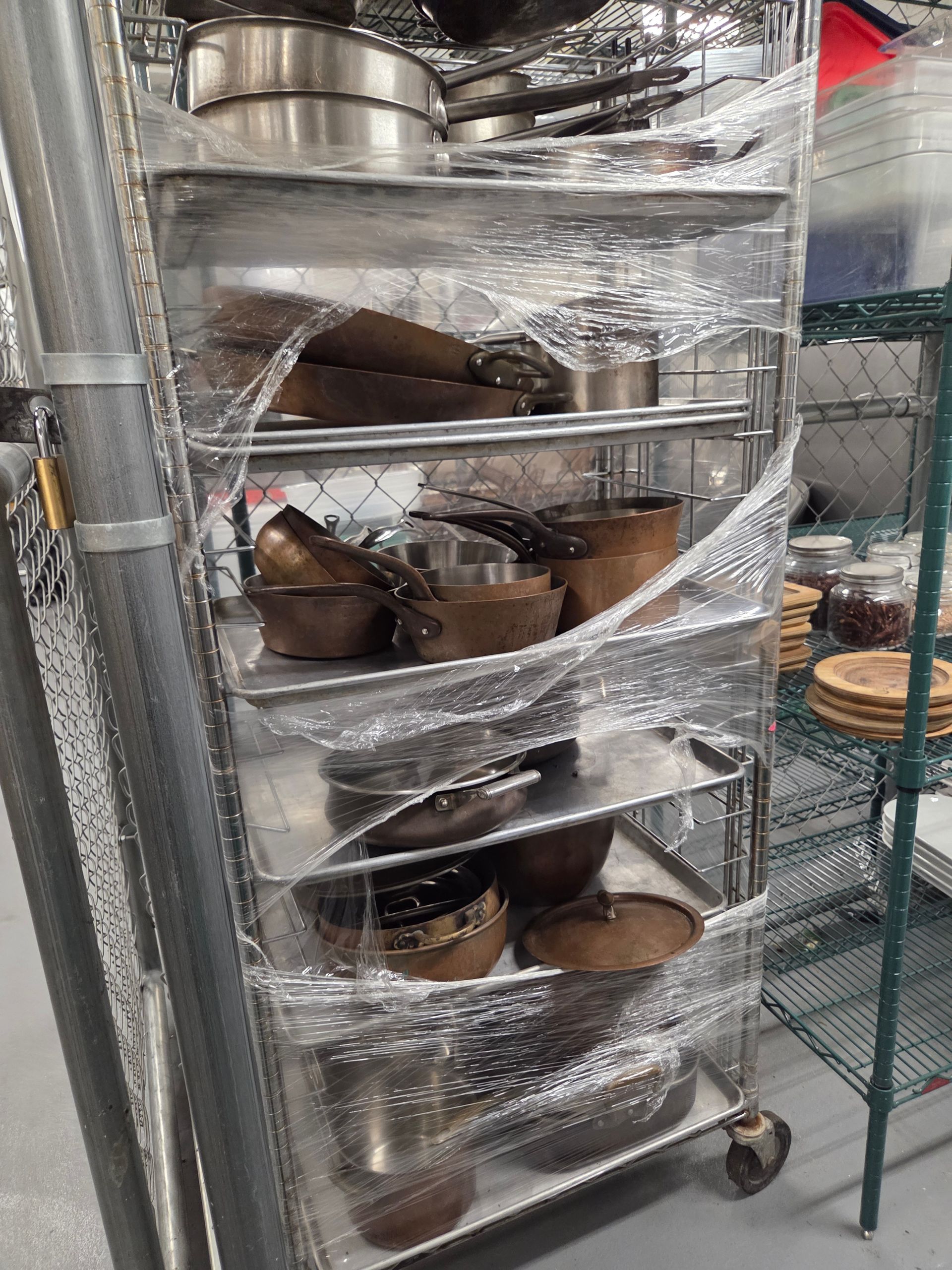 A stainless steel utility cart is wrapped in plastic and holding assorted cookware.