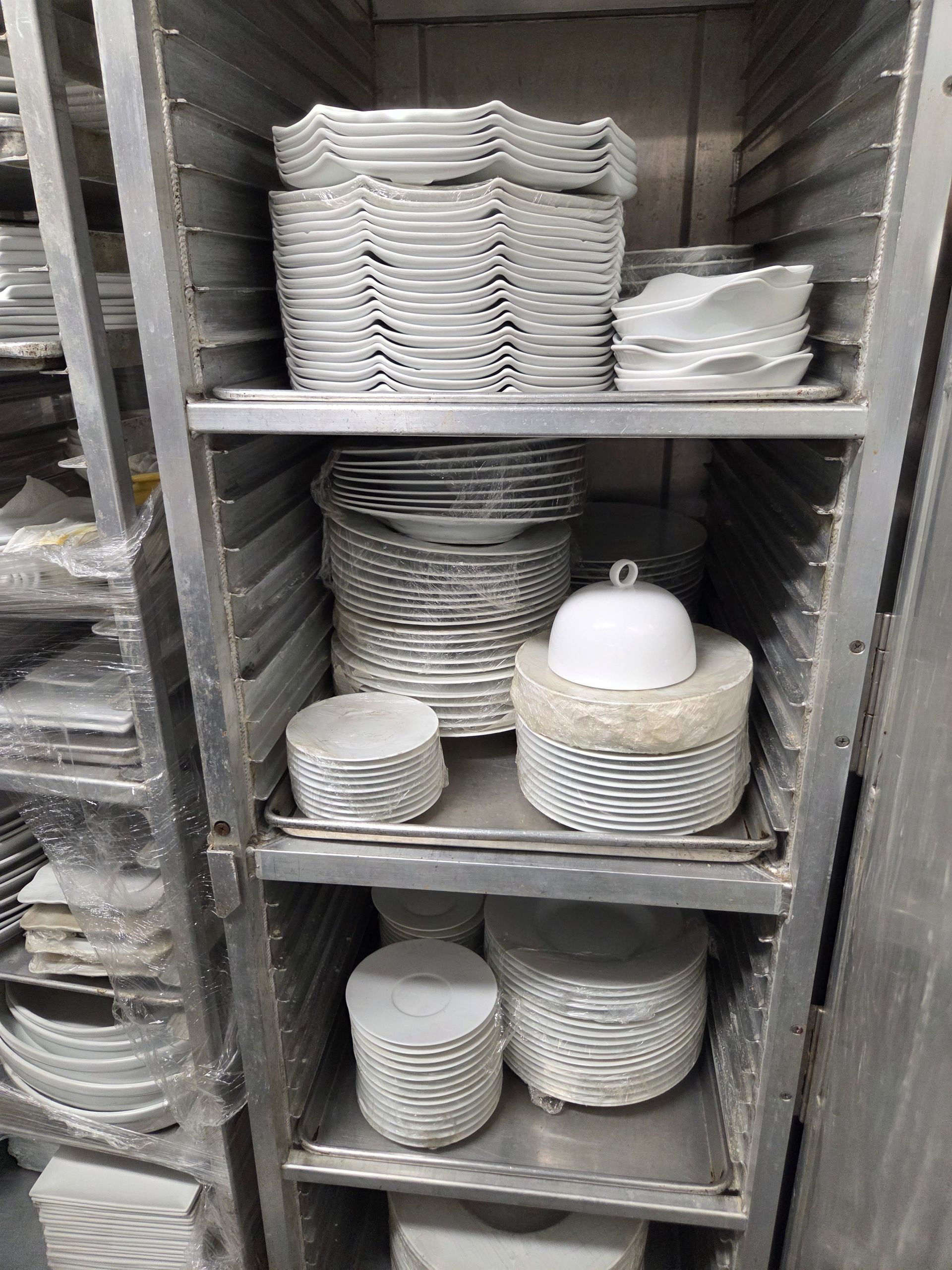 A stainless steel shelf unit filled with stacks of white ceramic plates and bowls in a kitchen.