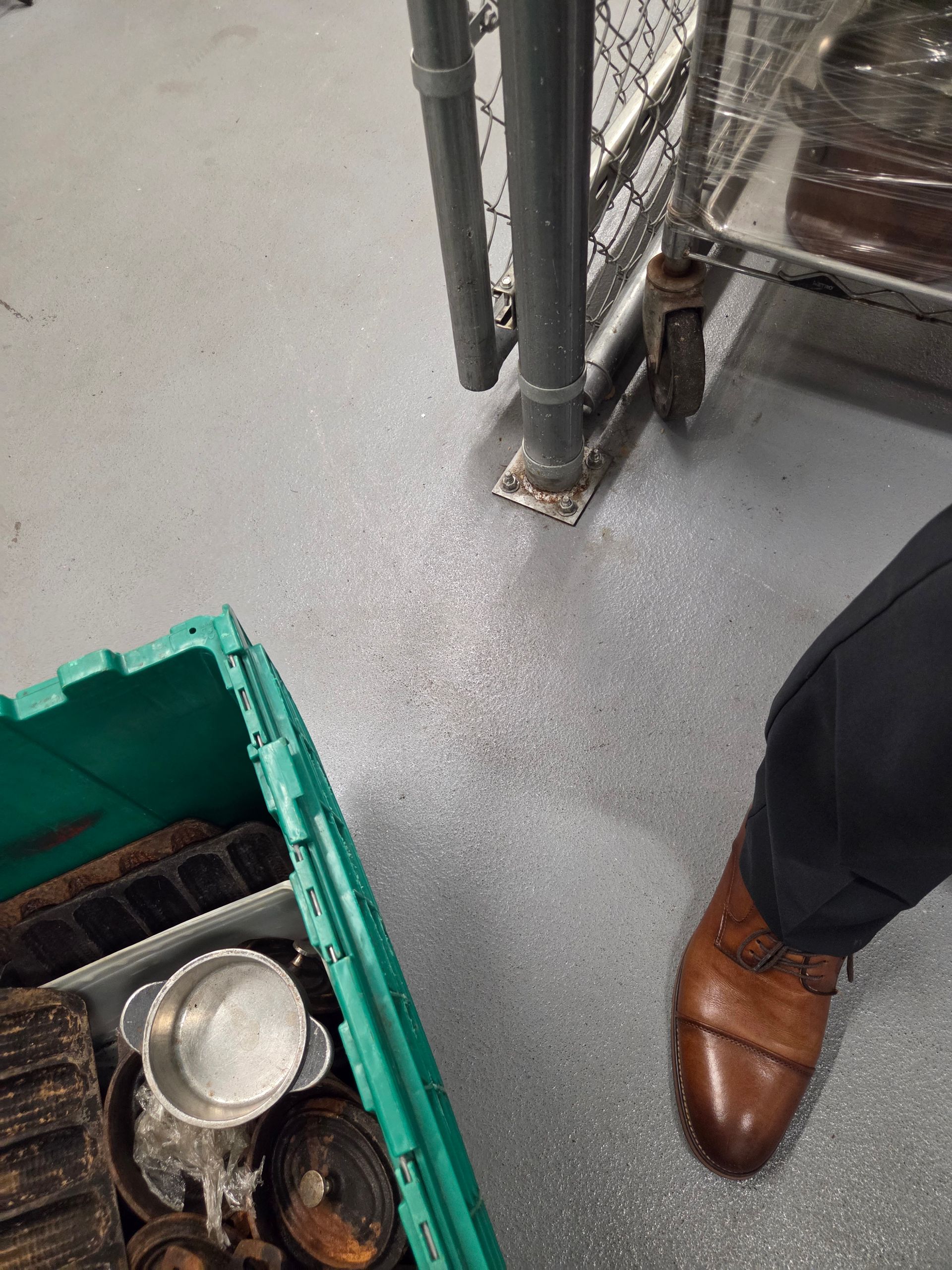 A person's brown shoe, green bin of objects, and metal cart on a gray floor.