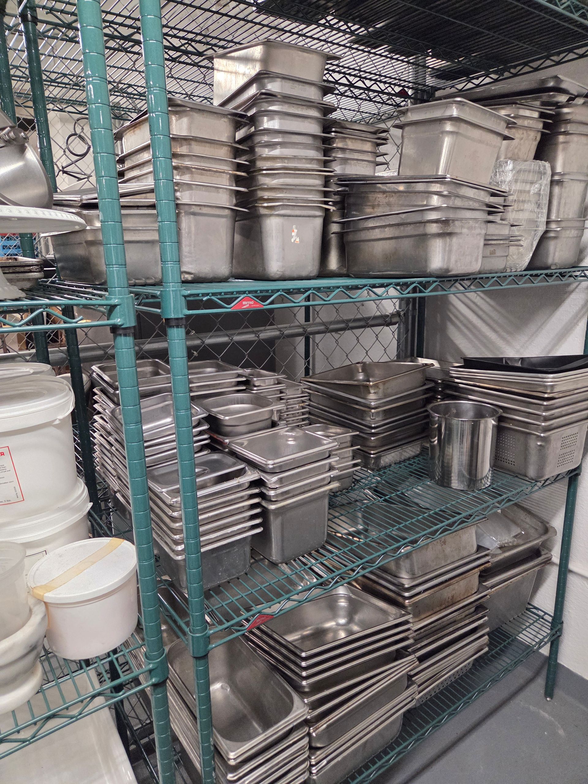 Shelving unit stacked with stainless steel food pans, pots, and containers in a commercial kitchen.