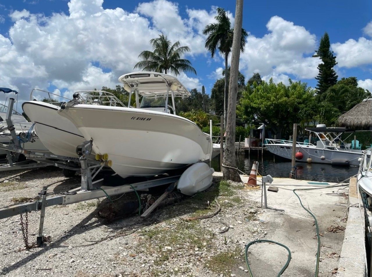 A white motorboat on a trailer, near a dock and palm trees, under a partly cloudy sky.