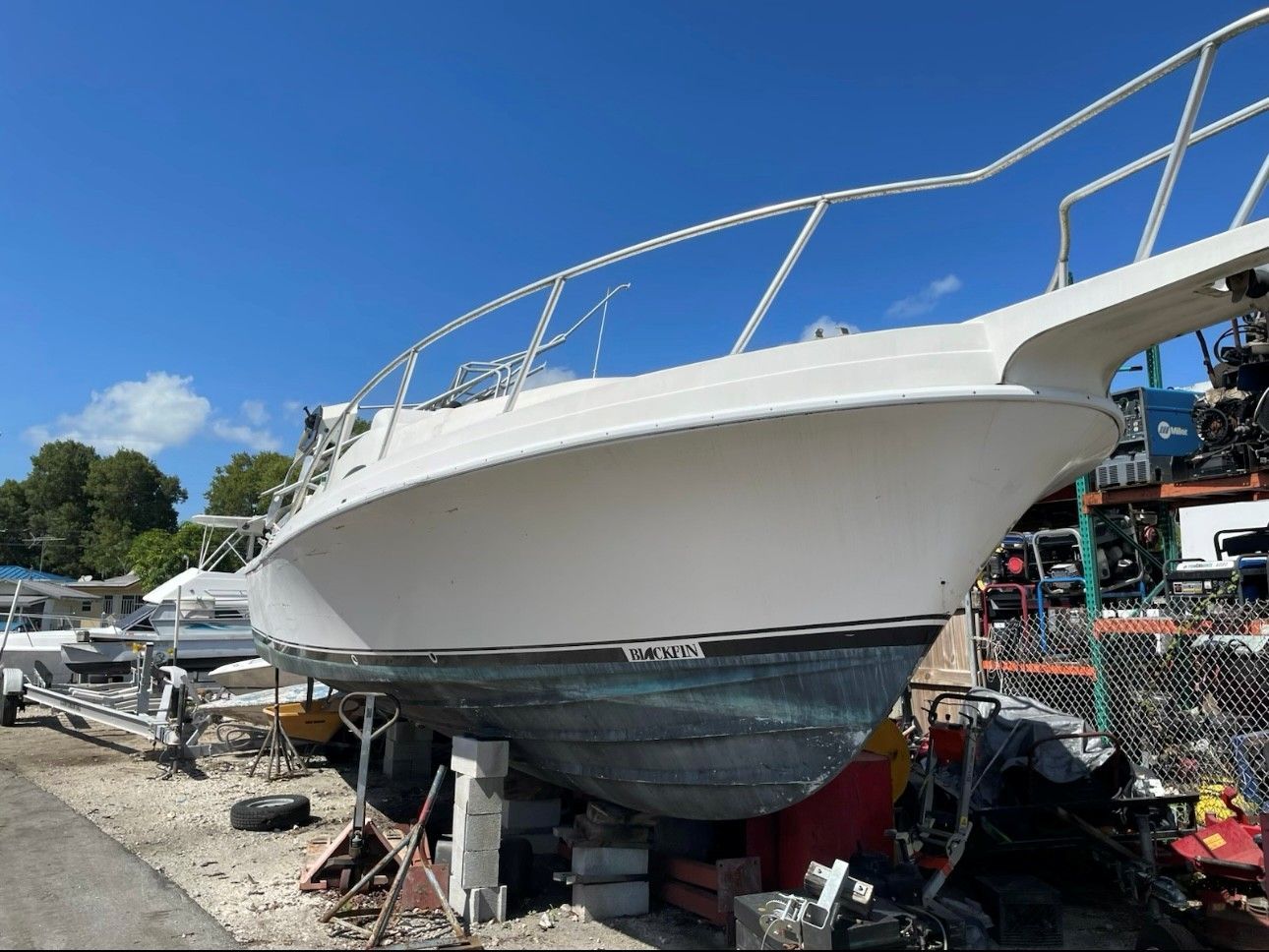White boat on blocks in a junkyard, blue sky overhead.