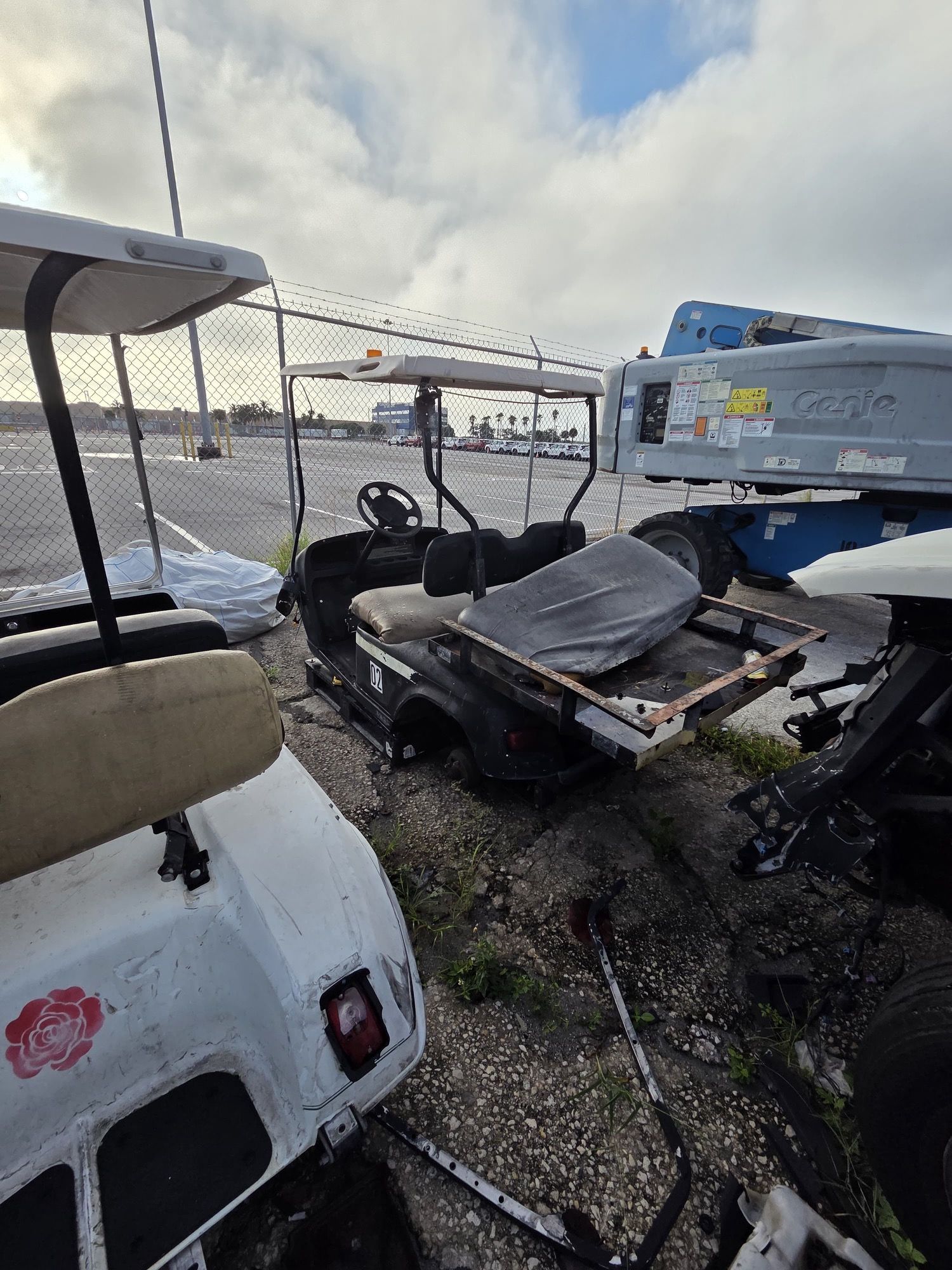 Golf carts in a lot near a fence. One covered with a sheet, one white, and a blue lift in the background.
