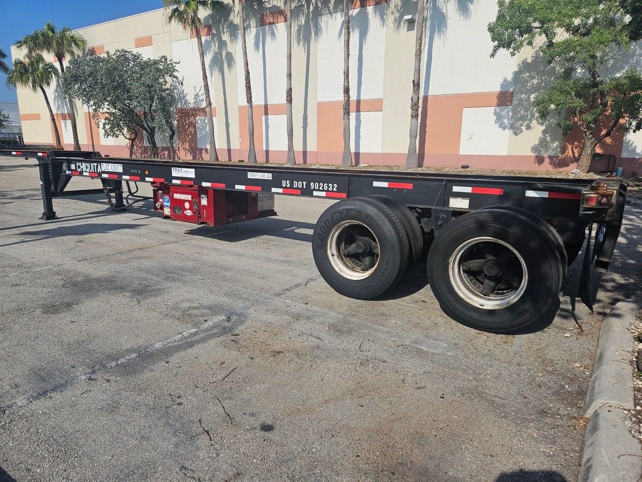 Empty black trailer on a gravel lot; red accents, tires, and a building with palm trees.