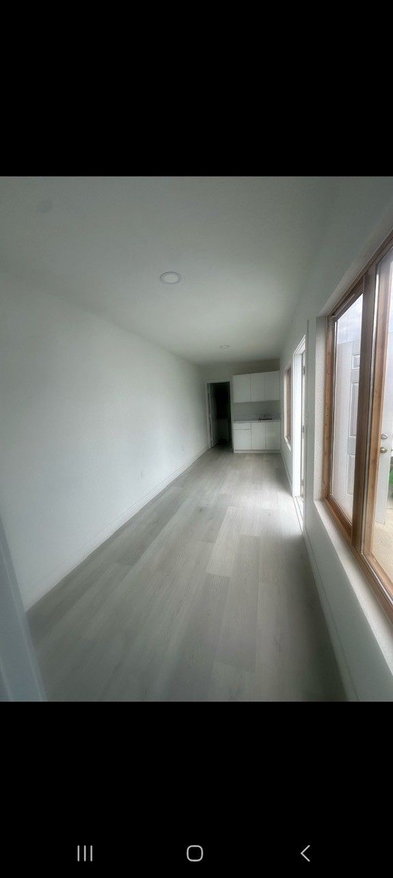 An empty, long, narrow room with white walls, wood-framed windows, and light gray flooring.