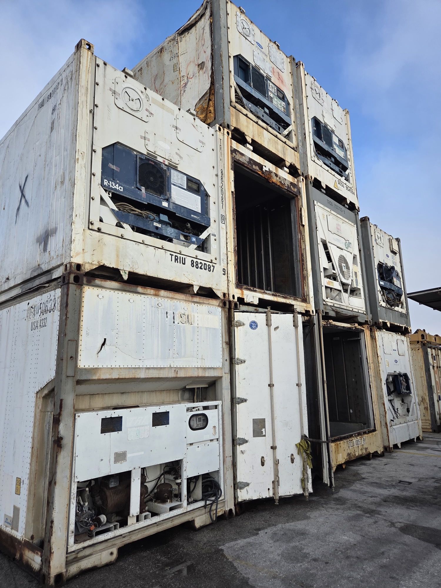Stacked white shipping containers, some open, showing refrigeration units, outdoors.