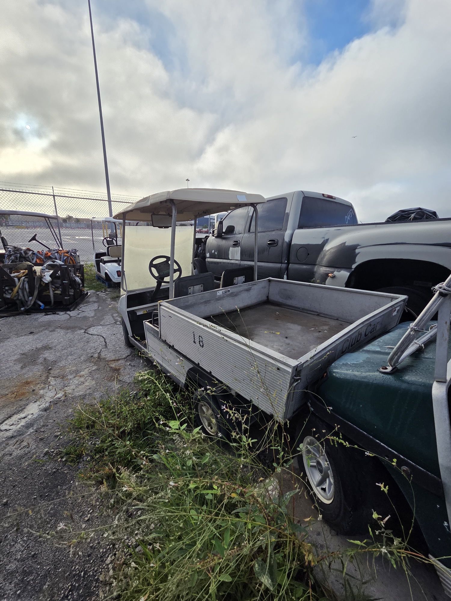 Gray utility golf cart with raised bed, parked next to a green golf cart, outside under cloudy sky.