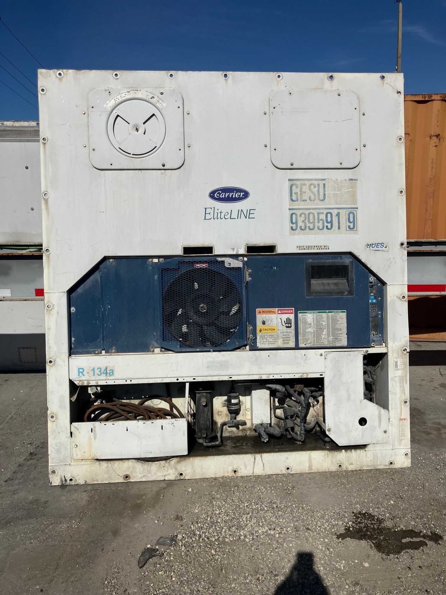 White and blue Carrier reefer unit with visible mechanical components, outdoors, sunny.