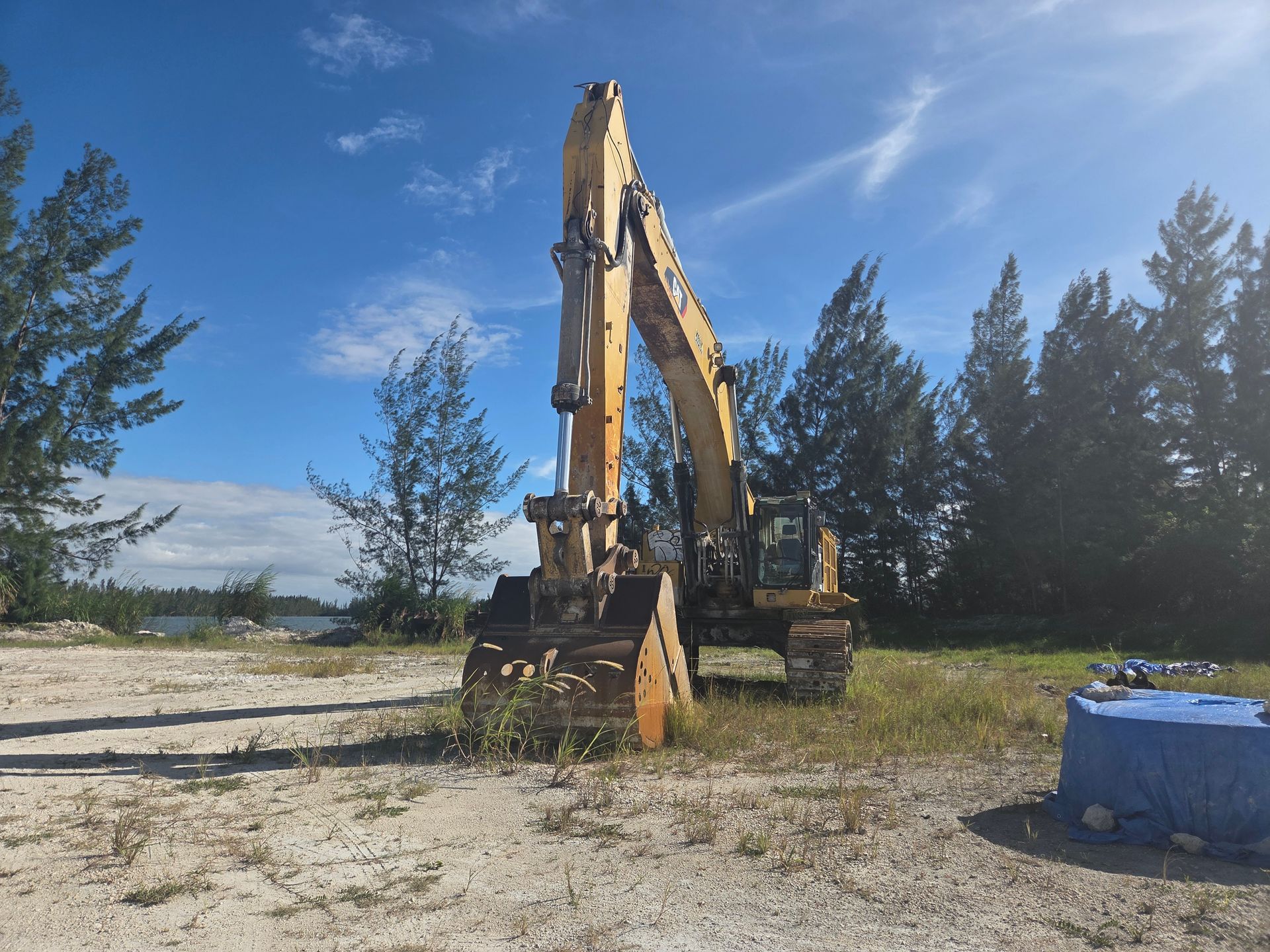 Yellow excavator on a dirt ground under a blue sky near trees and water.