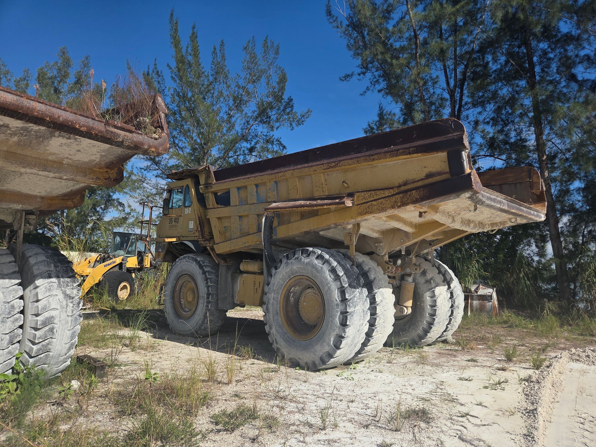Yellow dump truck with large tires in an outdoor setting. Another truck and trees are visible.