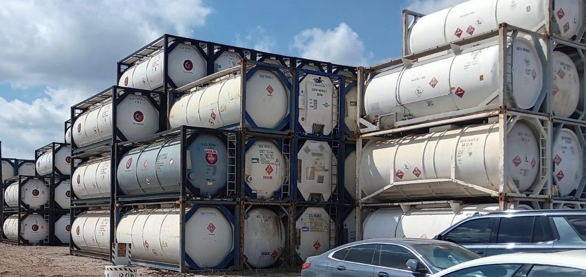 Stacks of white tanks in metal frames outdoors, some with hazard labels. Cars are in the foreground.