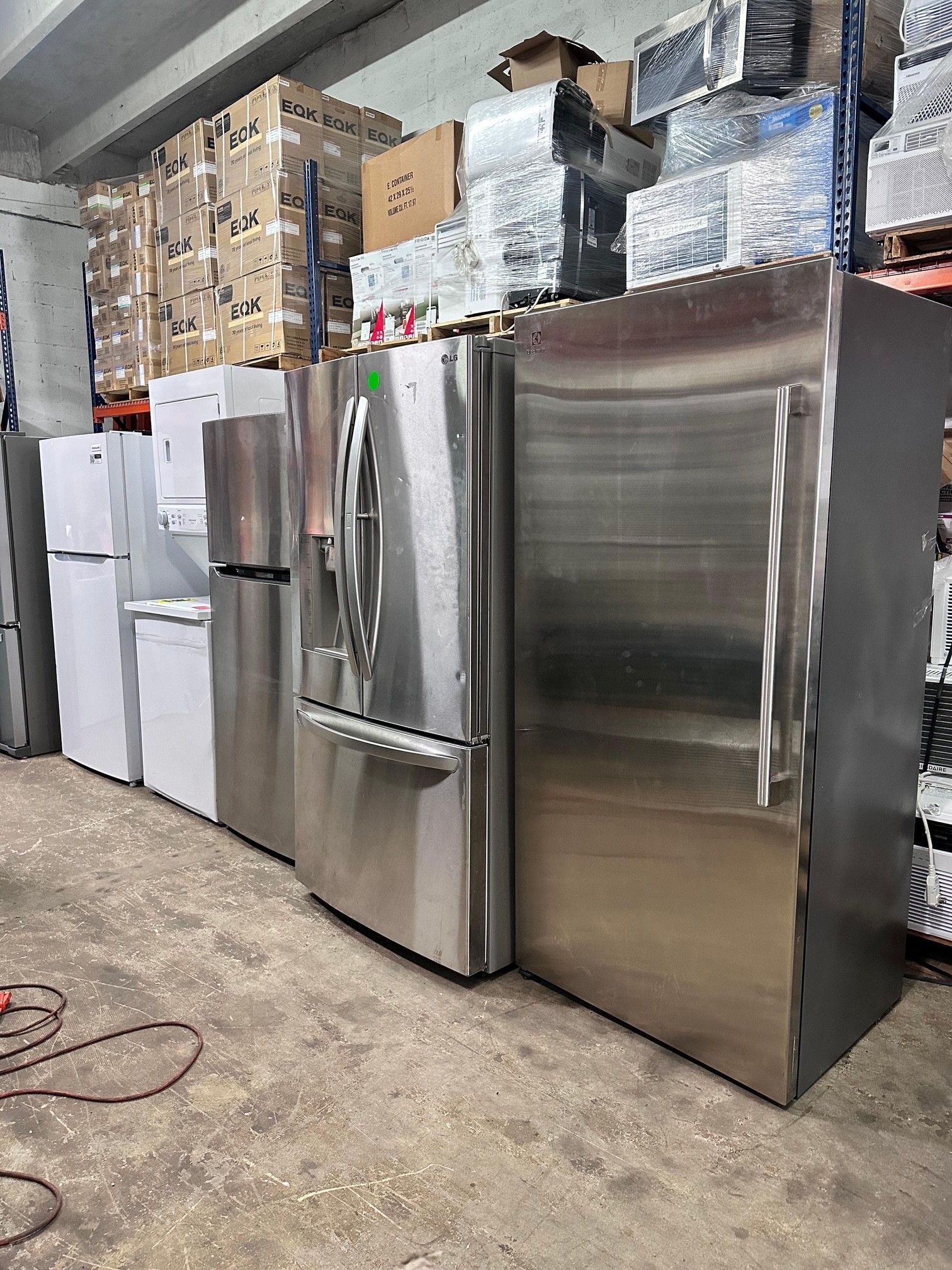 Refrigerators in a warehouse setting. Stainless steel and white refrigerators stand in a row. Shelves with boxes are in the background.