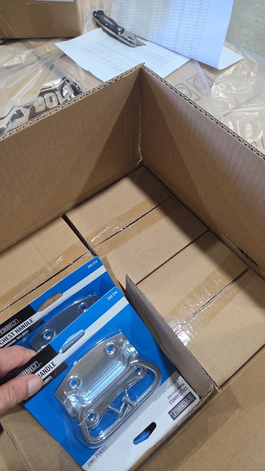 Hand holding a hardware package inside a cardboard box filled with more boxes in a warehouse setting.