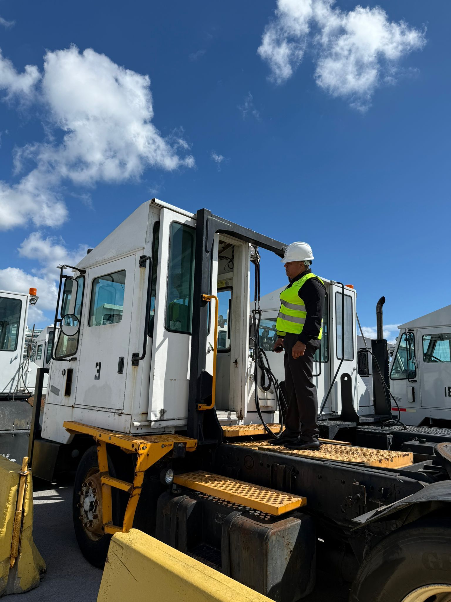 Man in a hard hat and vest on a white truck under a blue sky with clouds.