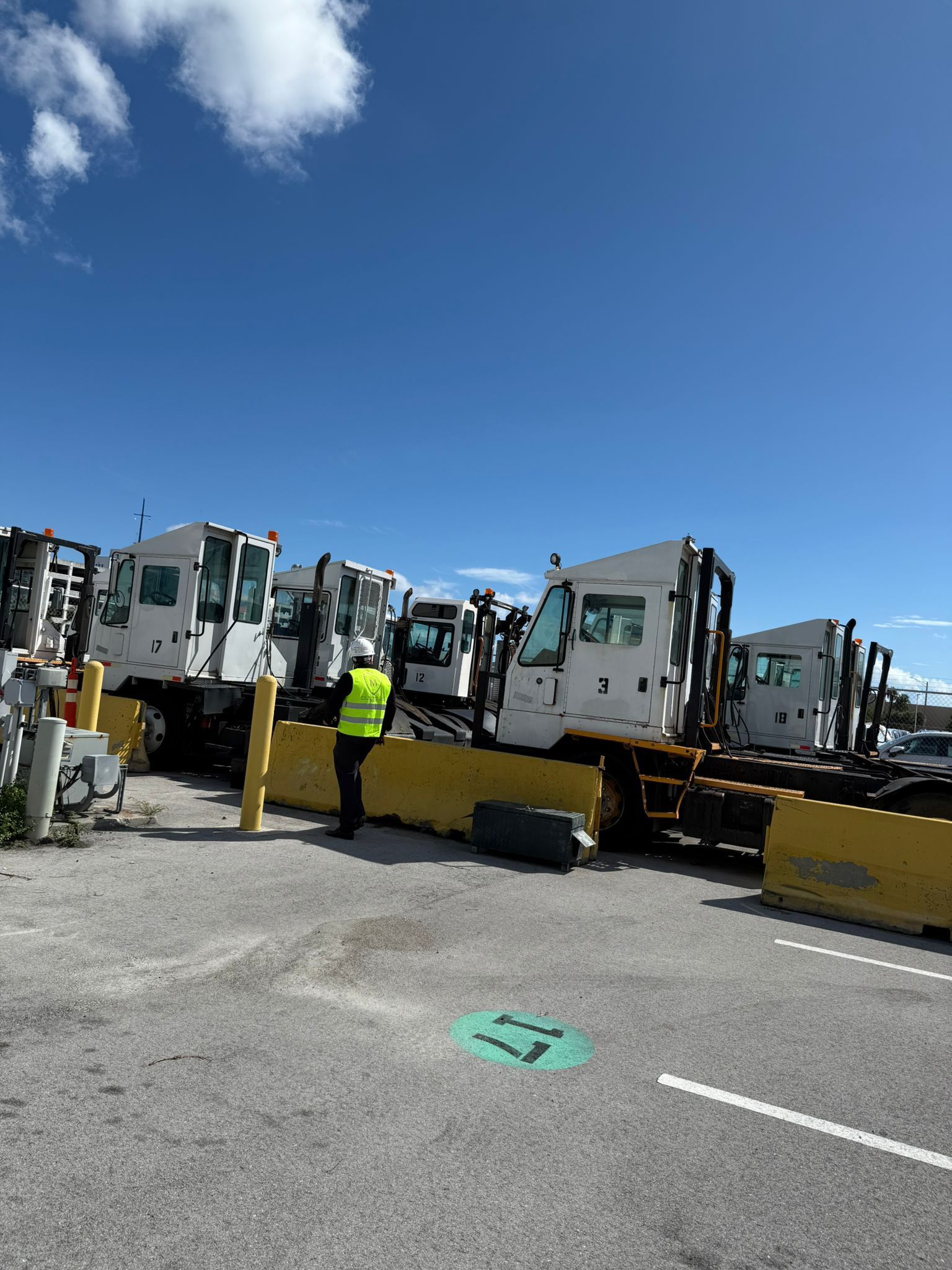 Person in a yellow vest stands near several white airport baggage tractors parked on a sunny day.