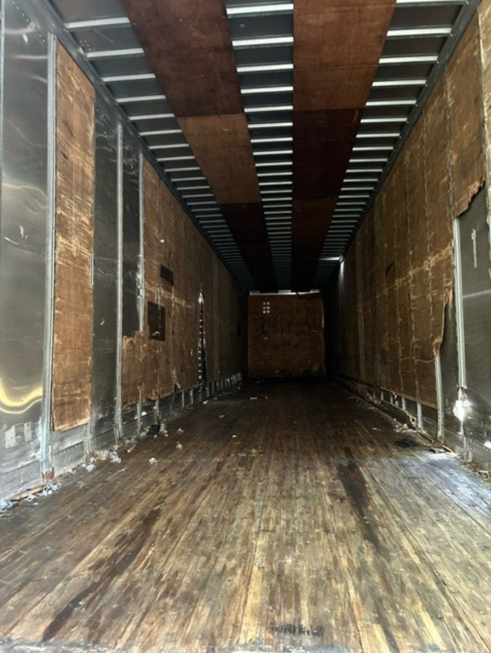 Interior of a dilapidated cargo truck, with peeling brown panels, wooden floor, and metal beams overhead.